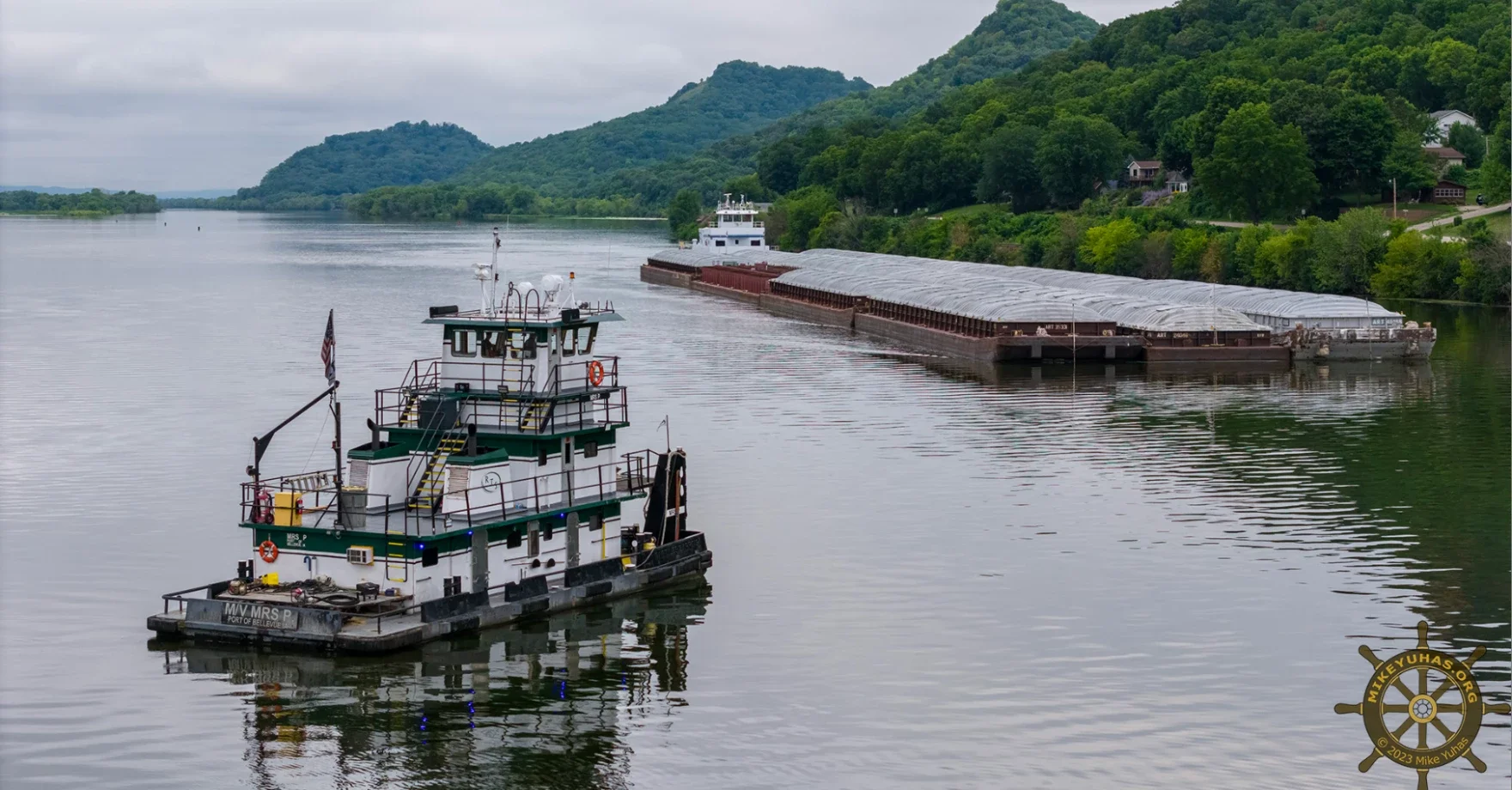 A towboat guides a long barge loaded with cargo along a calm river, with green hills and trees lining the shoreline under a cloudy sky.