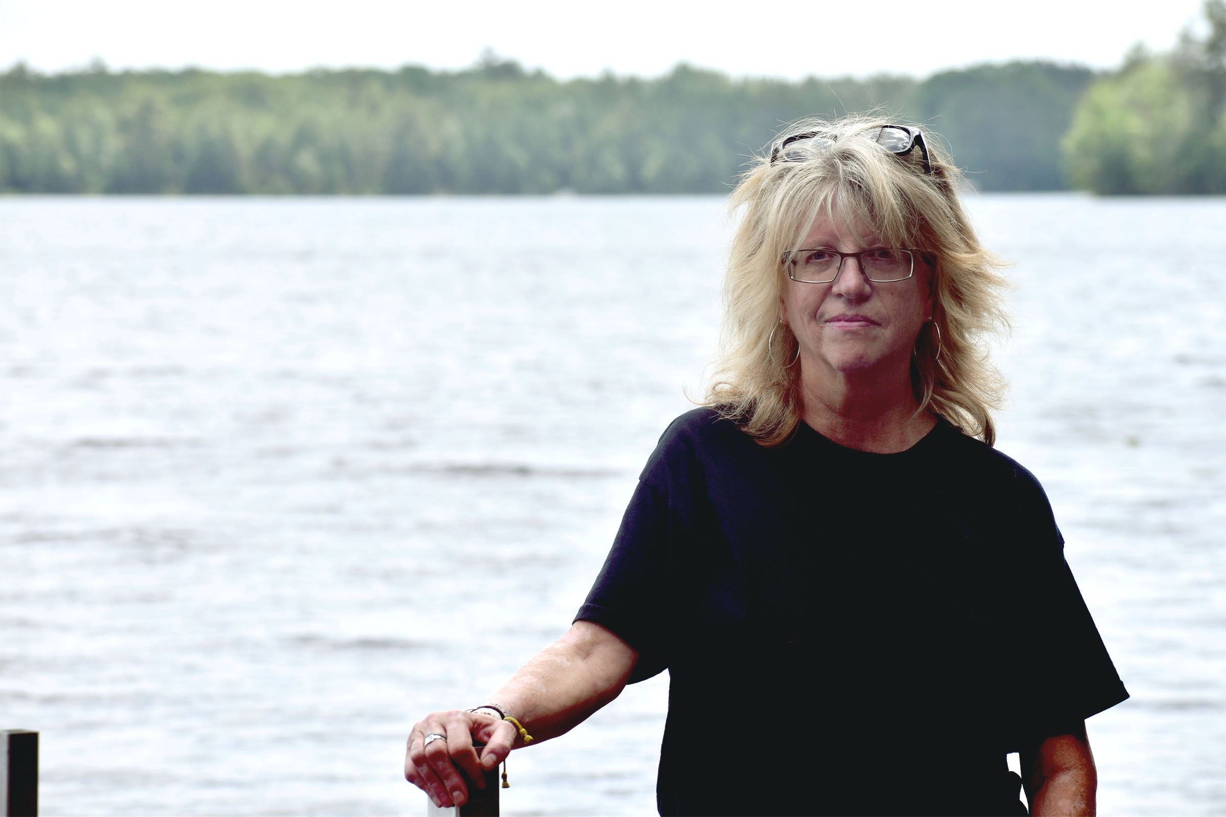 A woman with blond hair and glasses stands in front of a large body of water with a forested shoreline in the background.