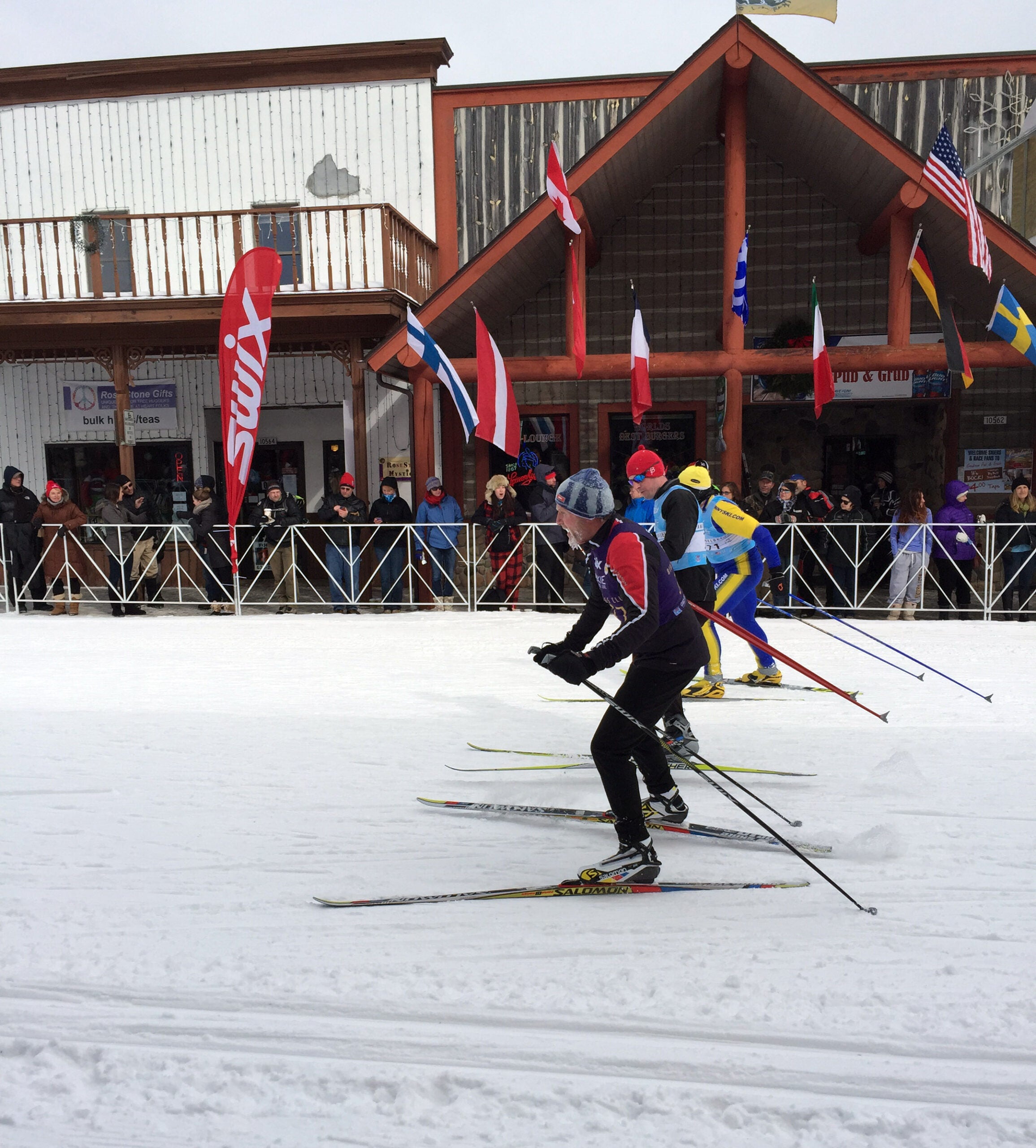 Three cross-country skiers race on a snowy course past a crowd and building with international flags displayed above.