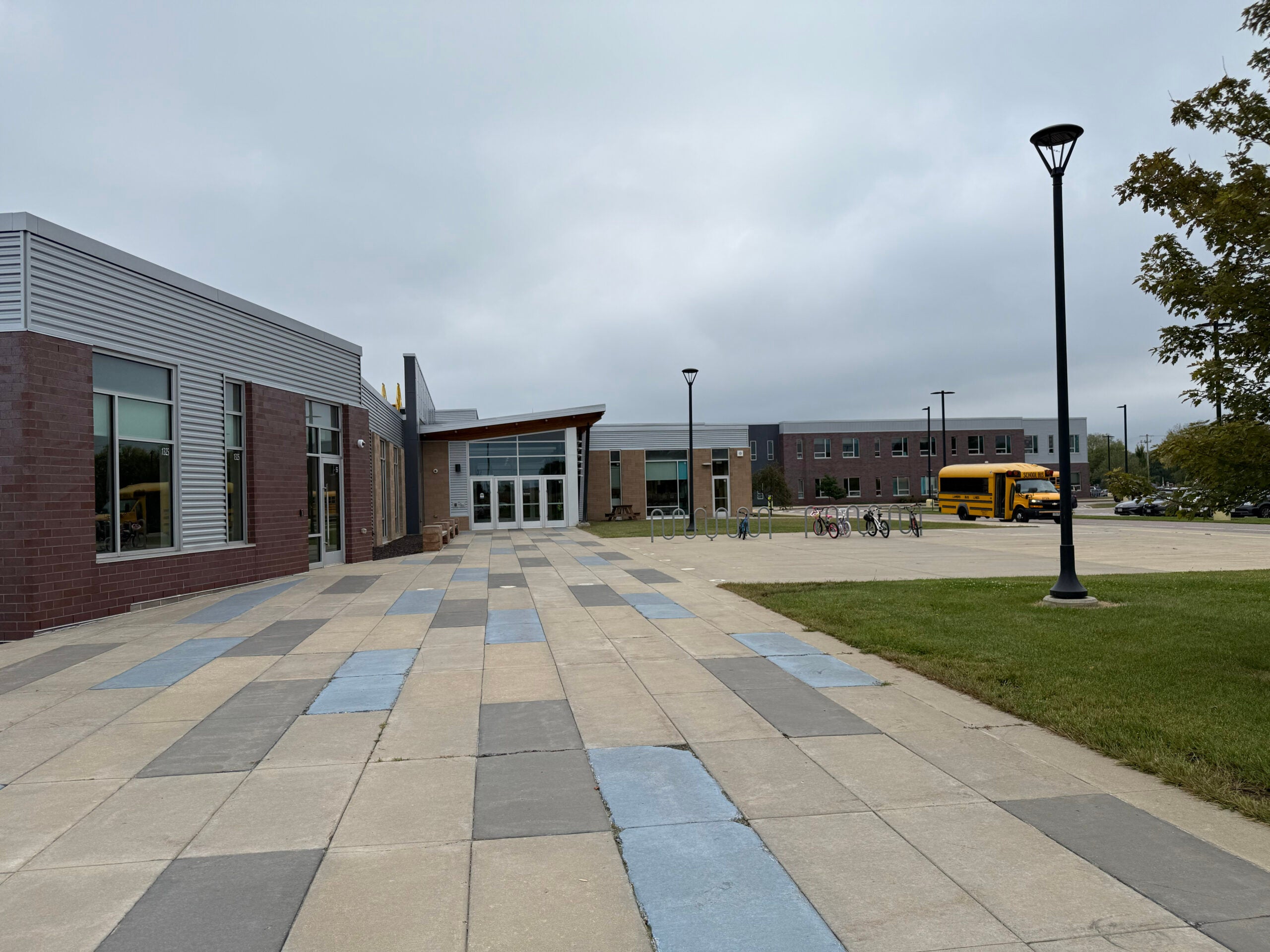 A modern school building with large windows and a checkered walkway; a yellow school bus is parked in the lot under a cloudy sky.