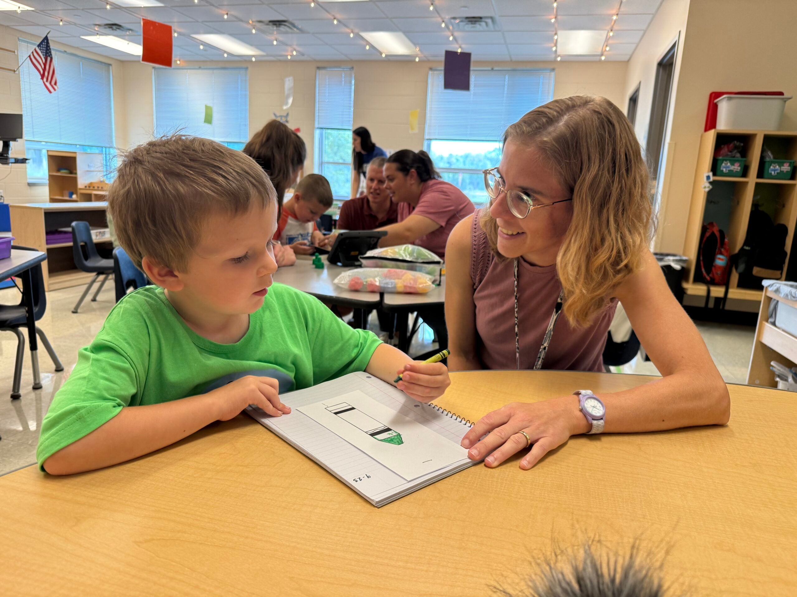 A young boy sits at a classroom table writing in a notebook while a teacher with glasses and a lanyard smiles and talks with him. Other students and adults are in the background.