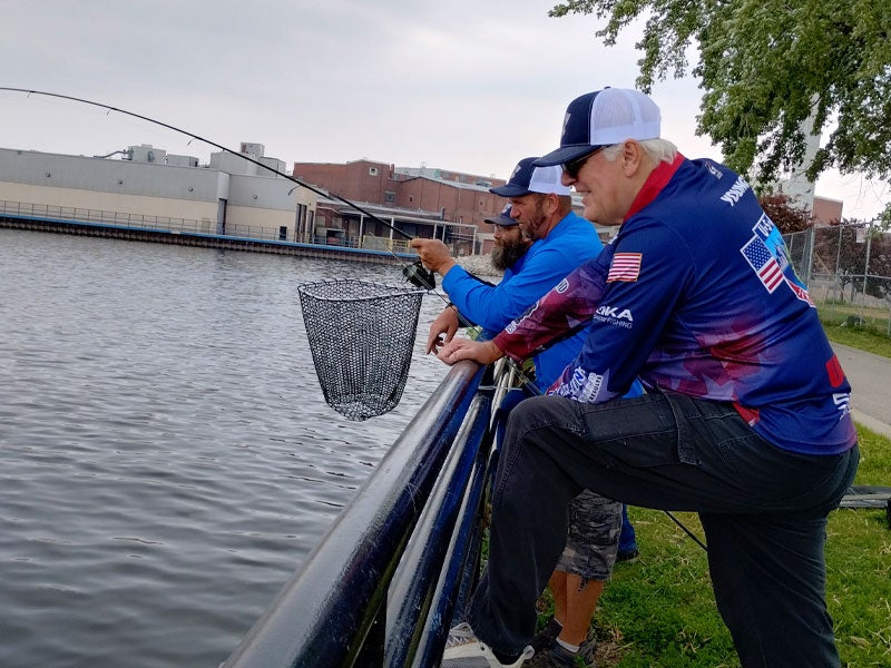 Two men stand by a railing near a body of water; one holds a fishing rod while the other holds a net over the water.