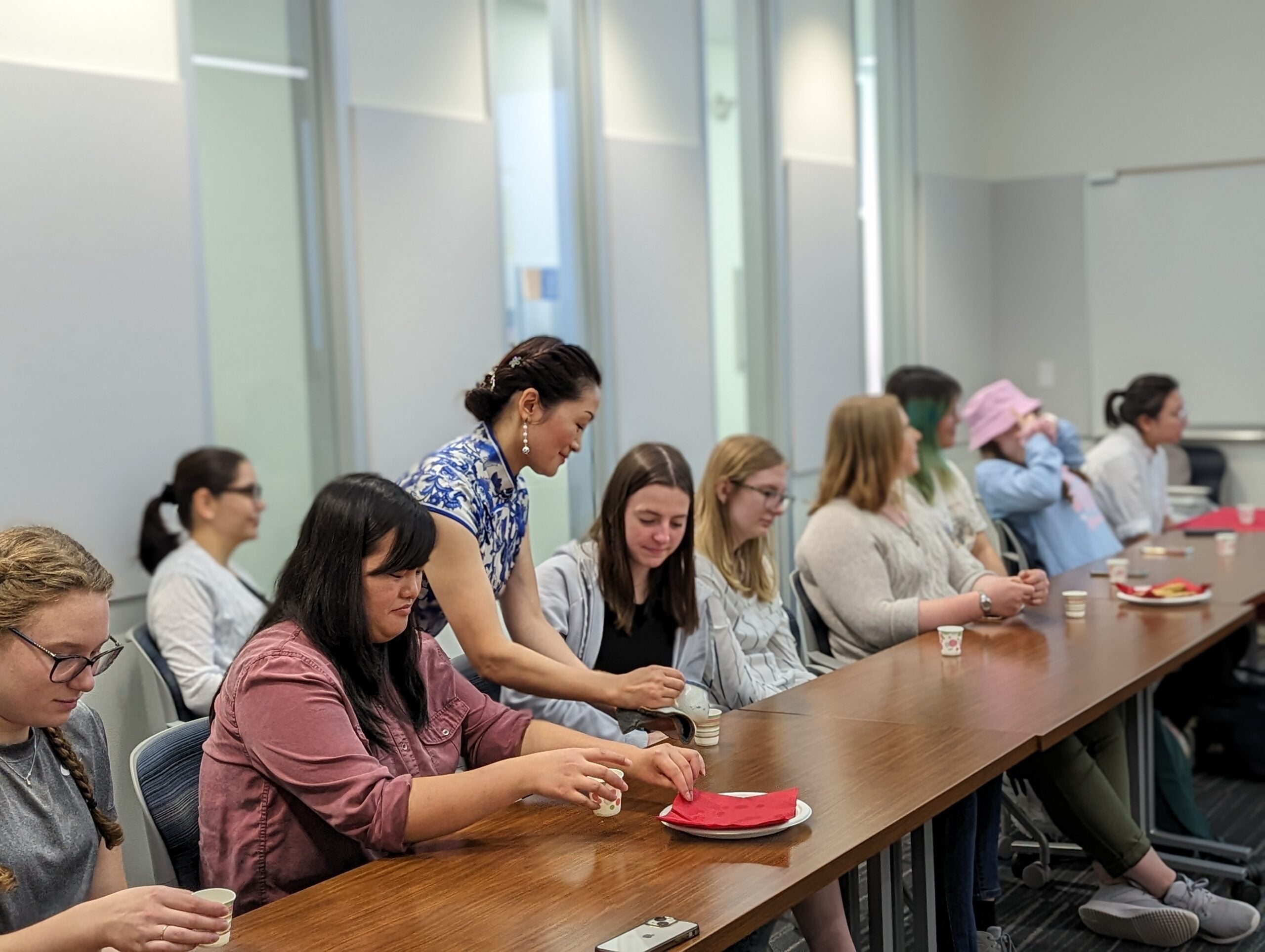 A group of people sitting at a long table in a classroom setting while a woman stands and assists one of them with a small cup.
