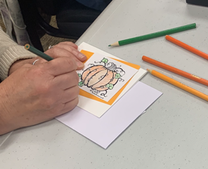A person colors a pumpkin-themed drawing on a card using colored pencils at a table.