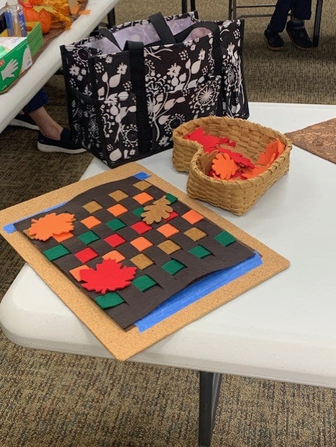 A table displays a woven craft made of brown, orange, and green strips with felt leaves, next to a basket of extra felt leaves and a patterned tote bag in the background.