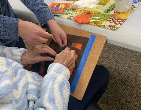 Two people work together on a craft project using colored felt on a board, with a puzzle and a cup on the table nearby.