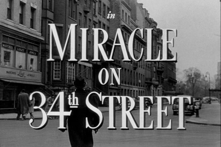 Black and white image showing the title Miracle on 34th Street over a city street scene with buildings and cars in the background.