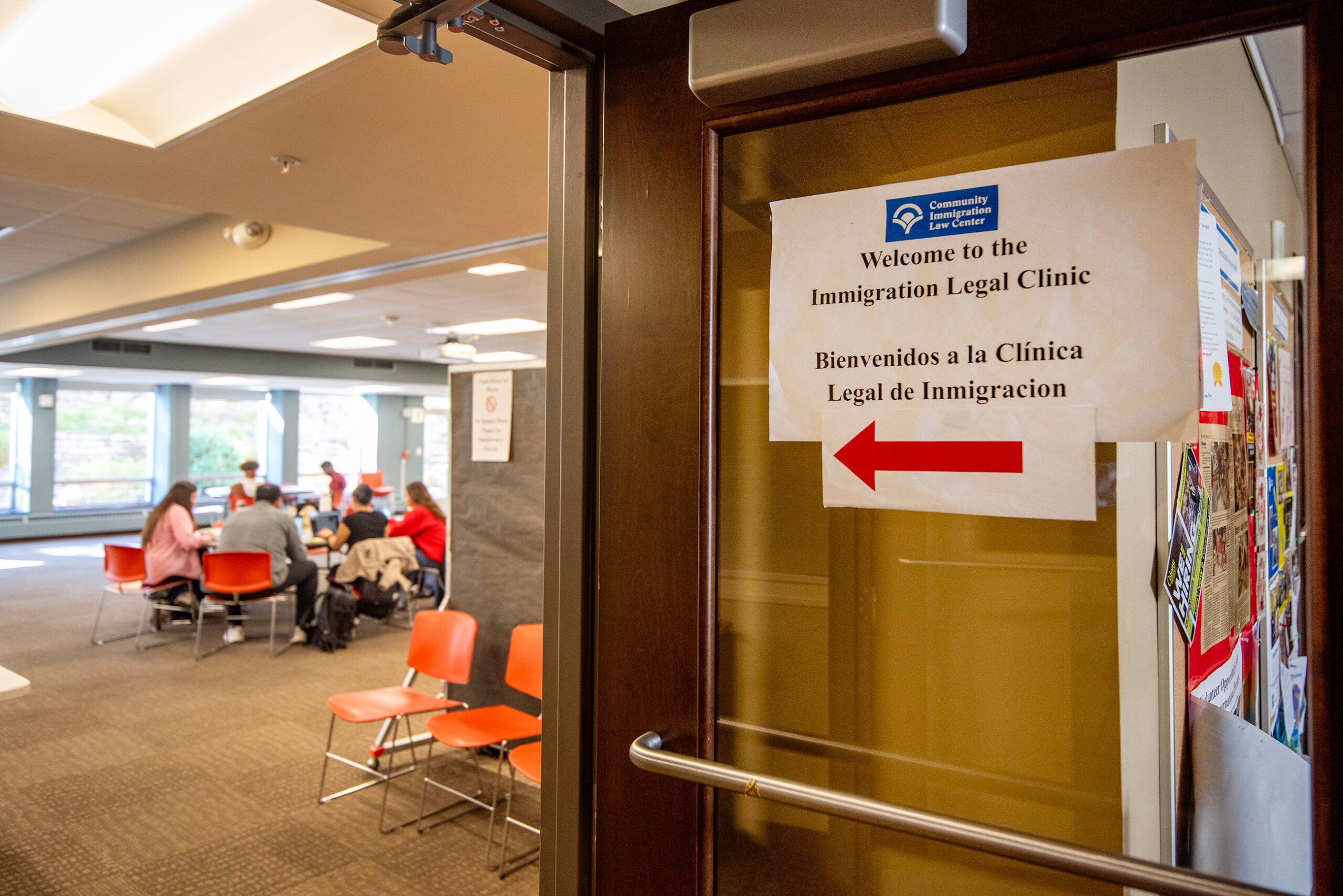 A sign on a door directs people to the Immigration Legal Clinic; inside, several people sit at tables in a meeting room.