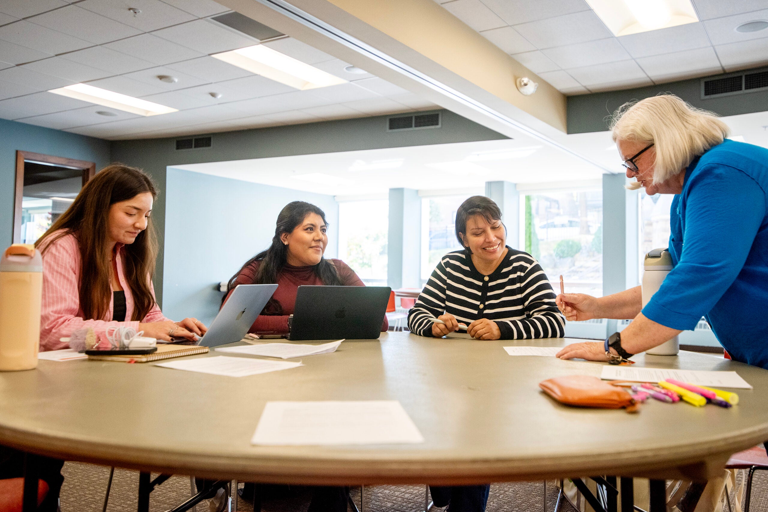 Four women are seated around a round table with papers and laptops, while an older woman stands and gestures during a discussion in a brightly lit room.