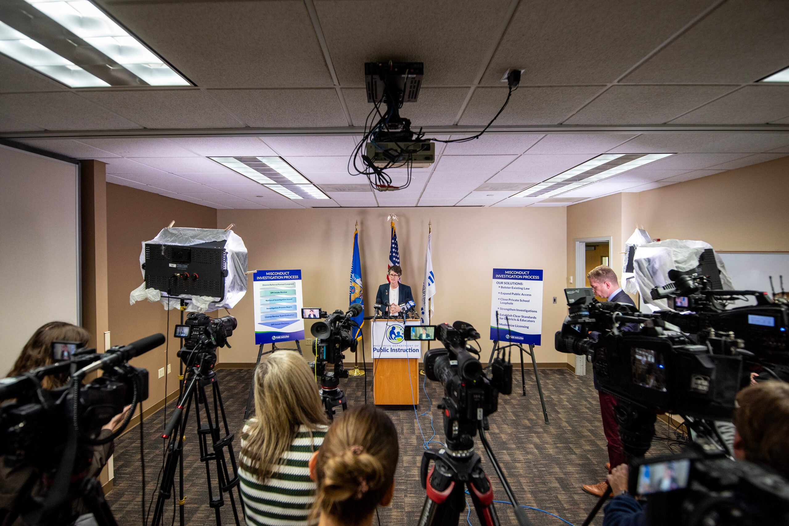 A person stands at a podium in a room, speaking at a press conference, surrounded by cameras, microphones, and information boards.