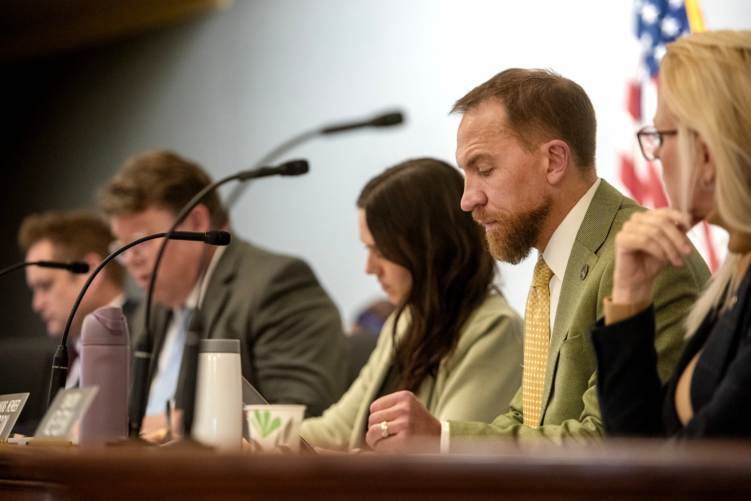 Five people sit in a row at a table with microphones, appearing focused during a formal meeting; an American flag is visible in the background.