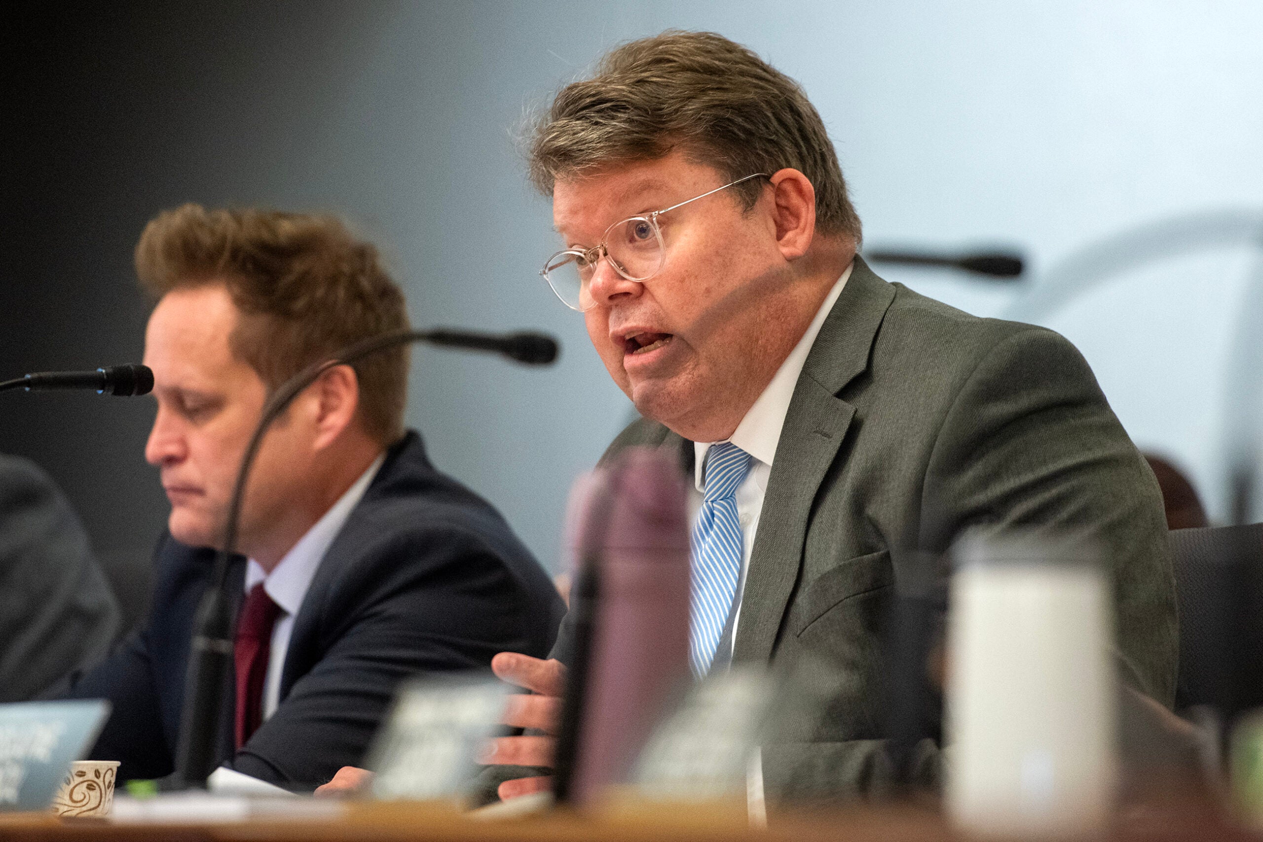 Two men in suits sit at a desk with microphones during a formal meeting or hearing. One man is speaking, while the other listens attentively.