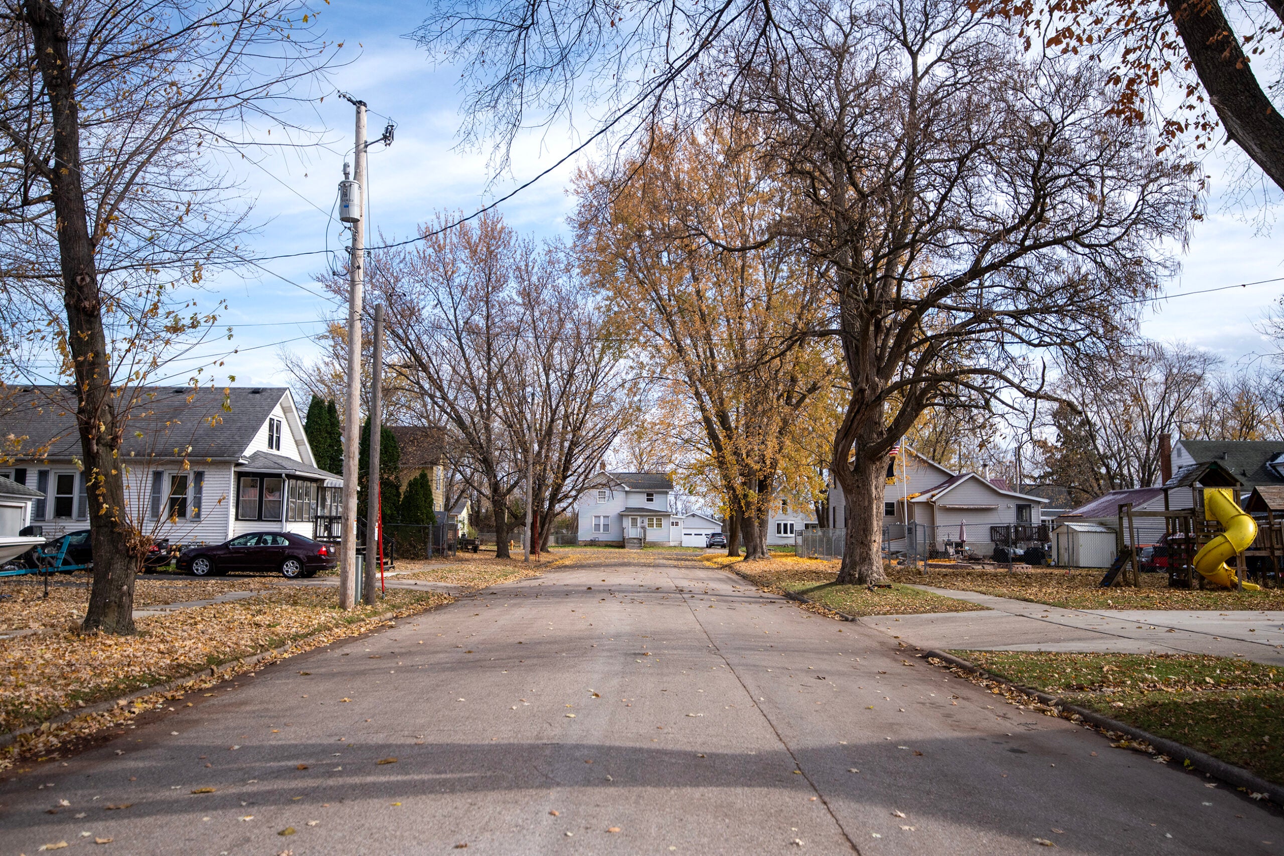 A quiet residential street lined with houses, leafless trees, and scattered autumn leaves; a playground with a yellow slide is visible on the right.