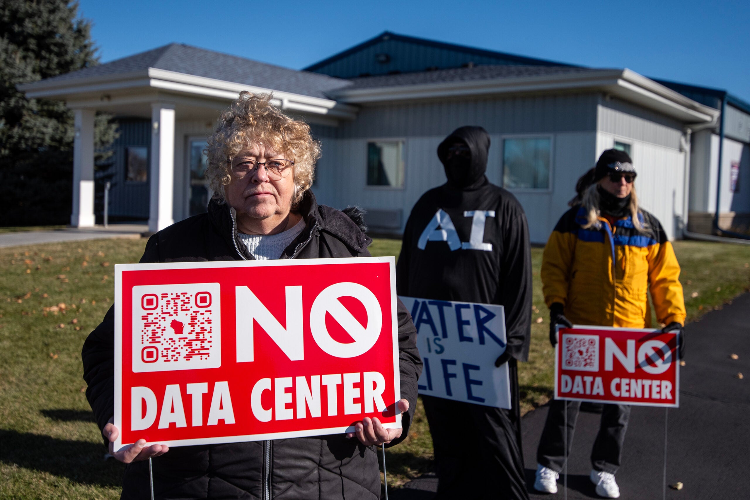 A woman holds a No Data Center sign, while two other people stand nearby with protest signs in front of a building.
