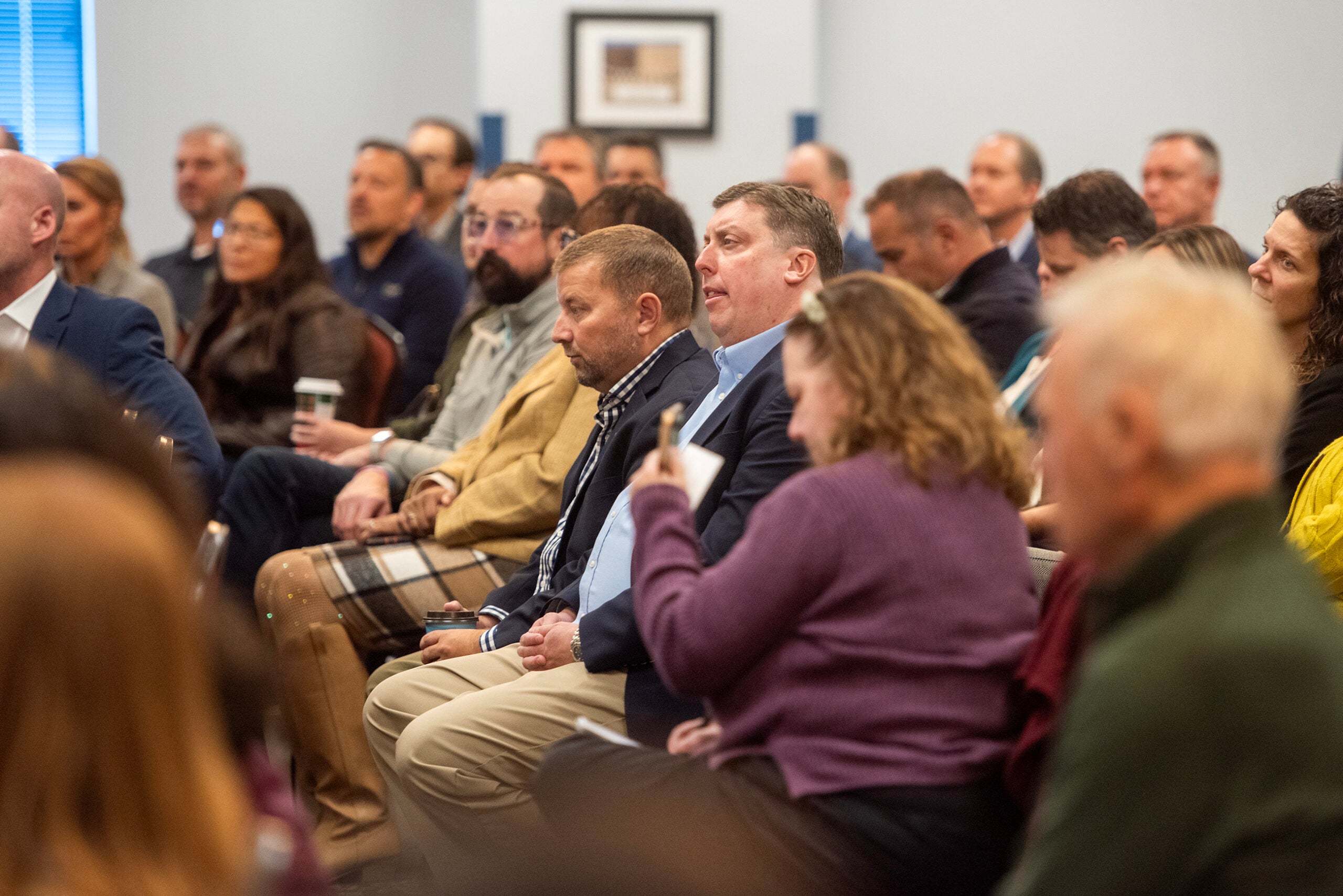 A group of adults sits in rows, attentively listening during an indoor meeting or presentation.
