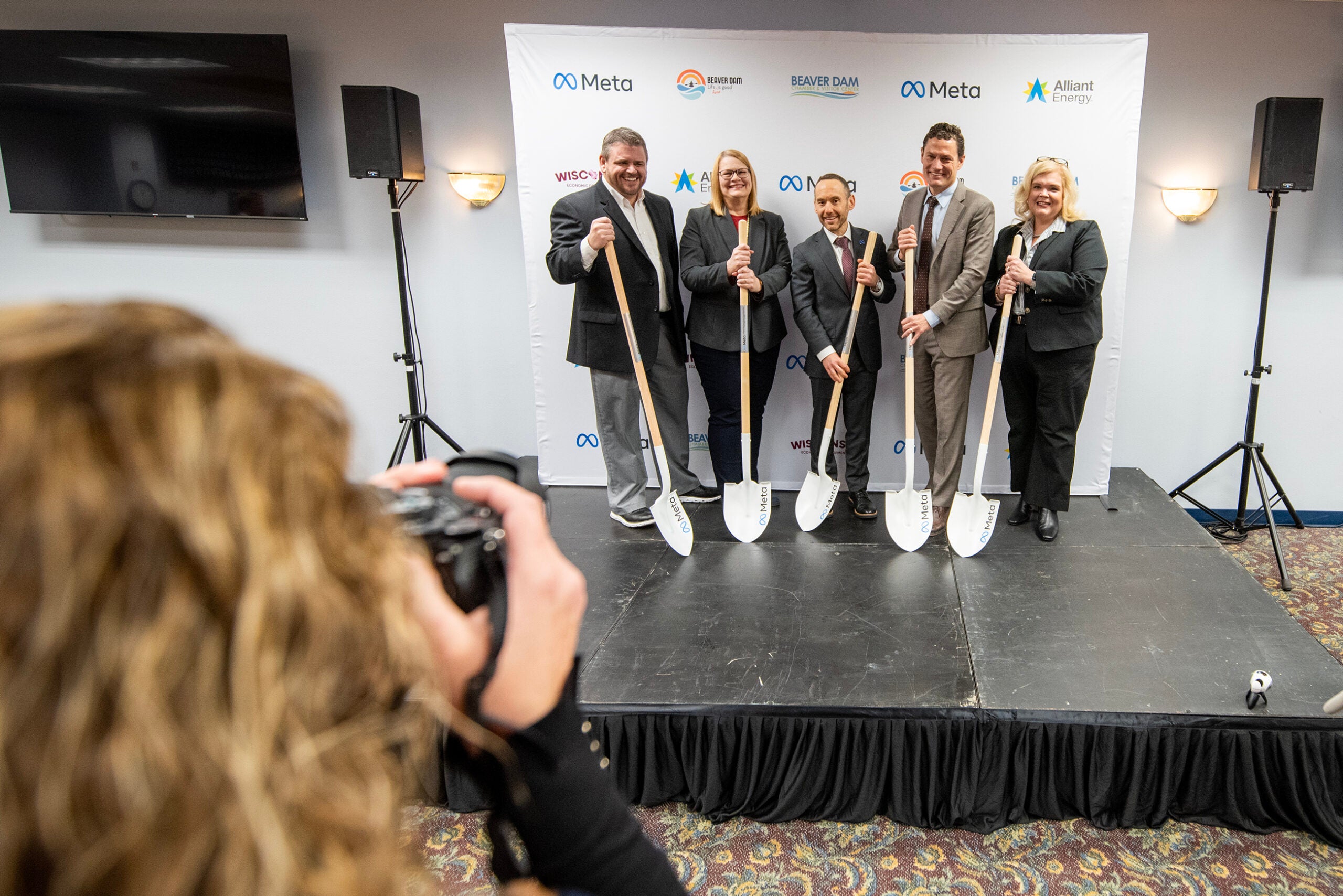 Five people in business attire stand on a stage holding ceremonial shovels, posing for a photo at a groundbreaking event in front of a Meta-branded backdrop.