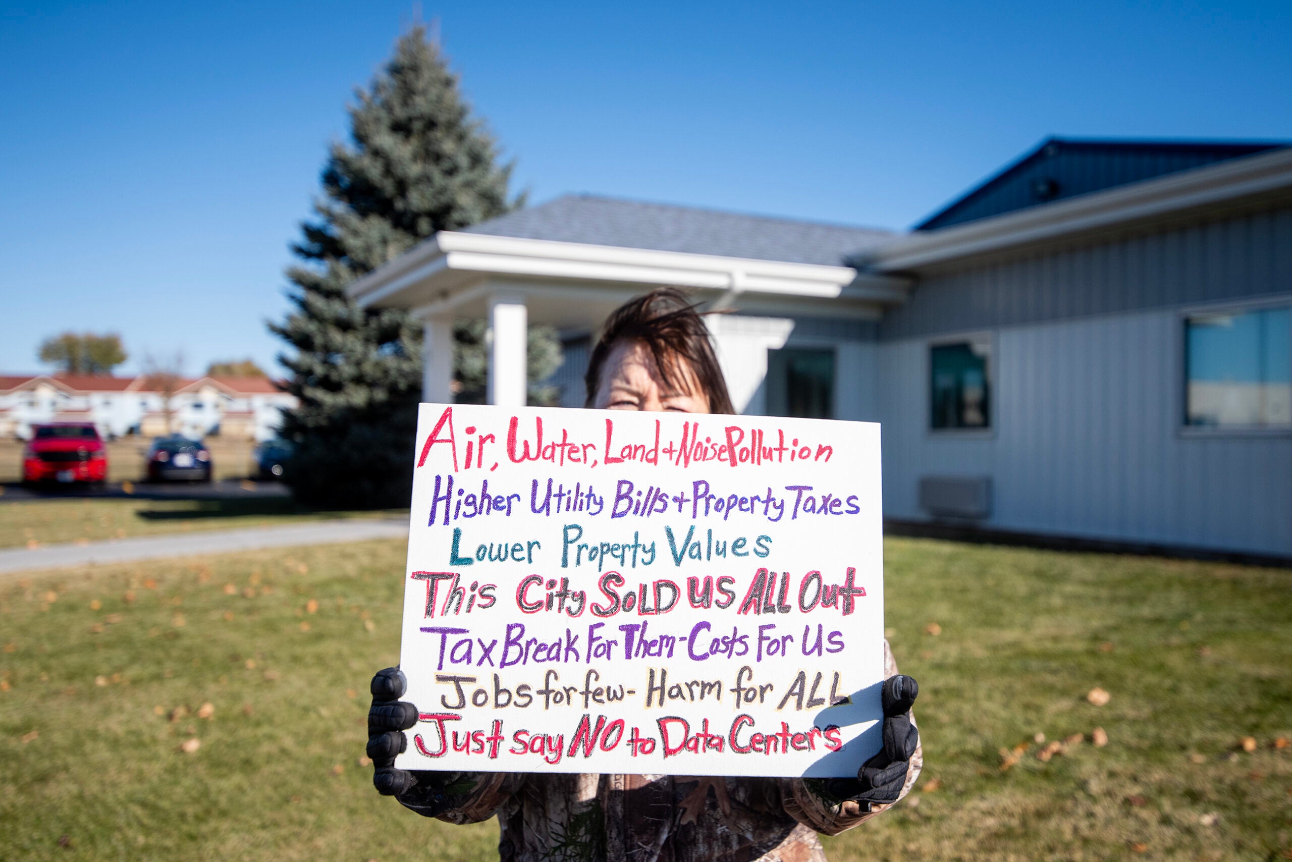 A person stands outside holding a sign protesting data centers, citing concerns about pollution, higher costs, lower property values, and tax breaks for companies.