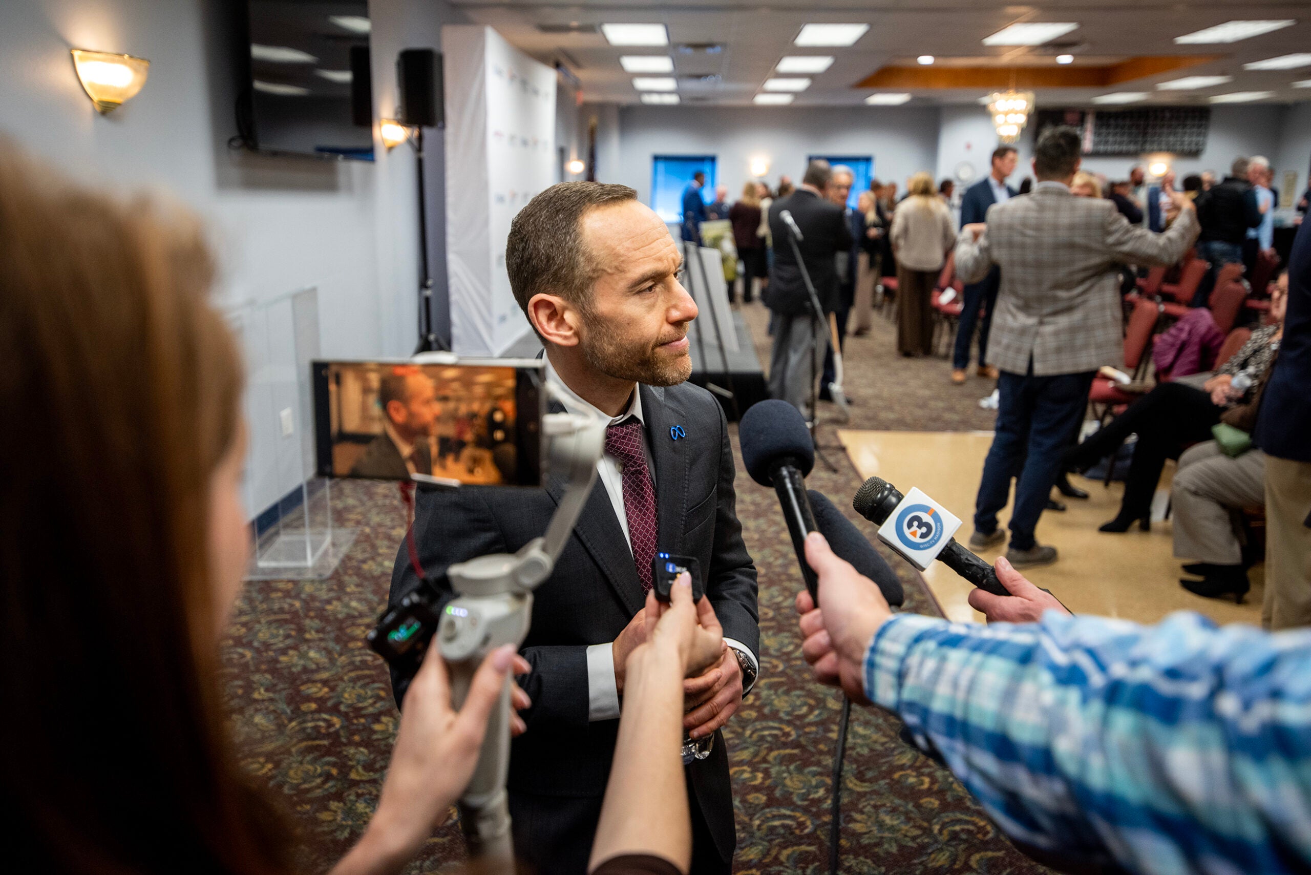 A man in a suit speaks to reporters holding microphones and recording devices at an indoor event with people in the background.