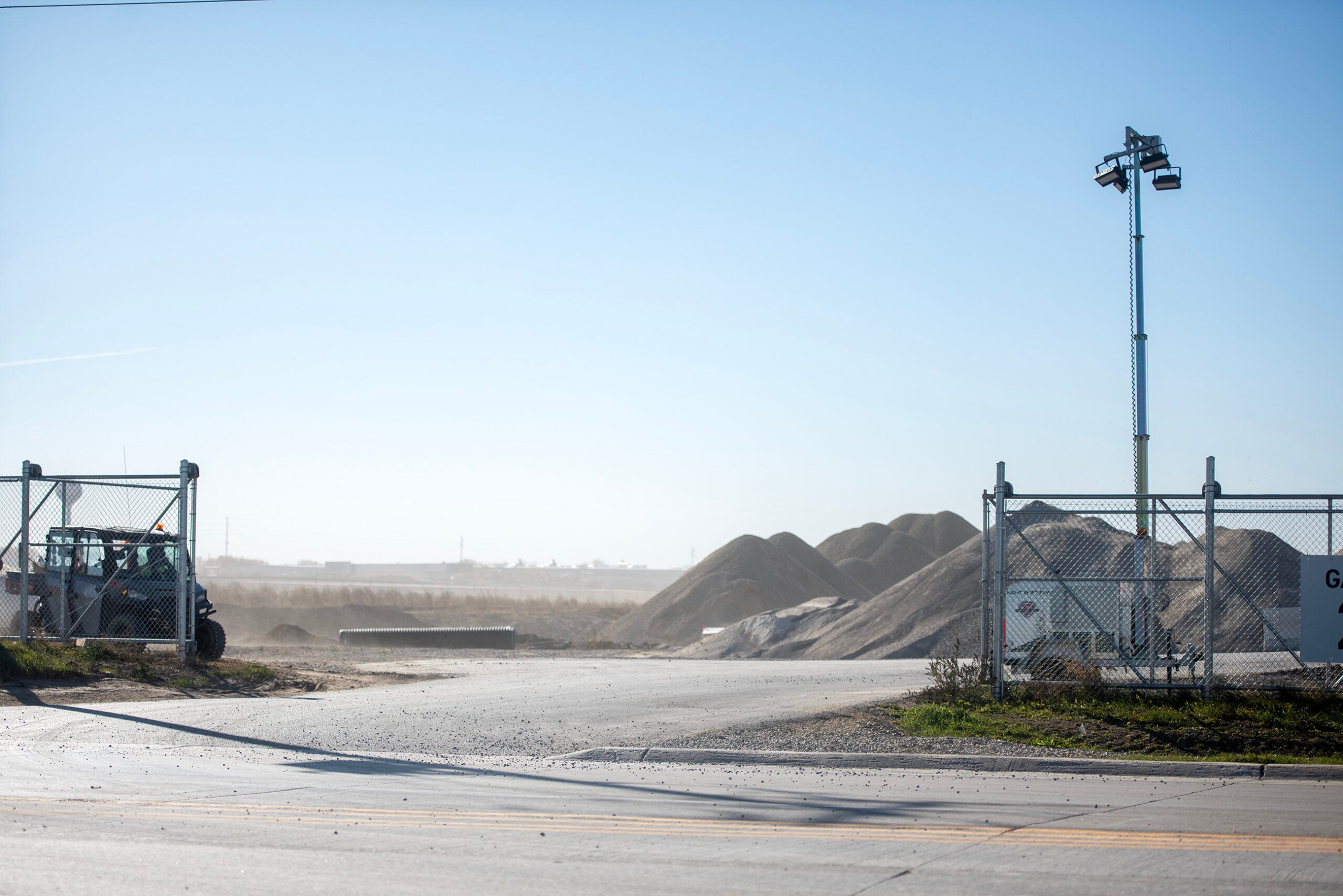 A fenced industrial yard with piles of gravel, construction equipment, and a tall floodlight pole under a clear blue sky.