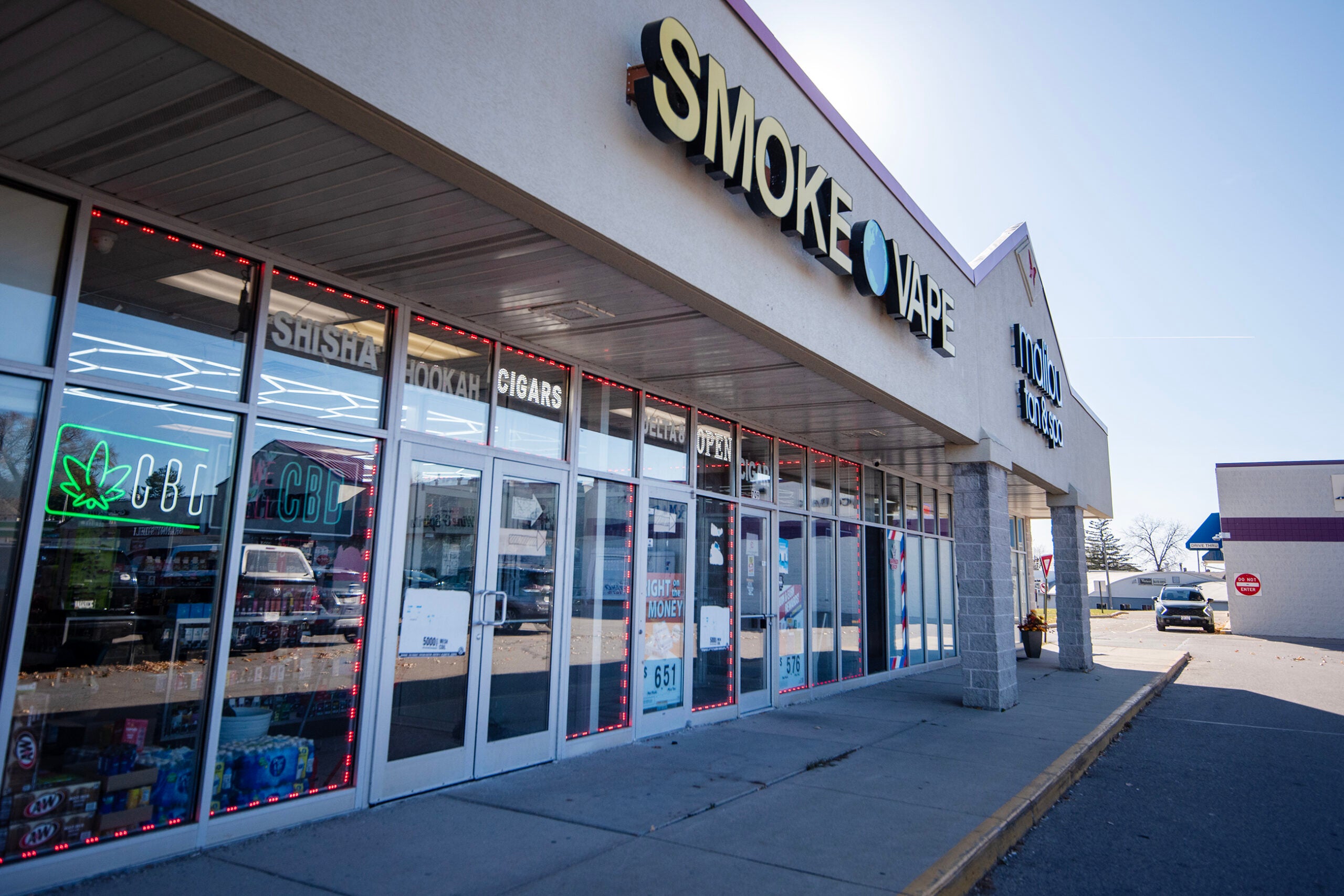 A strip mall storefront with large Smoke Vape sign, glass windows displaying products, and neon signs for CBD and tobacco items.