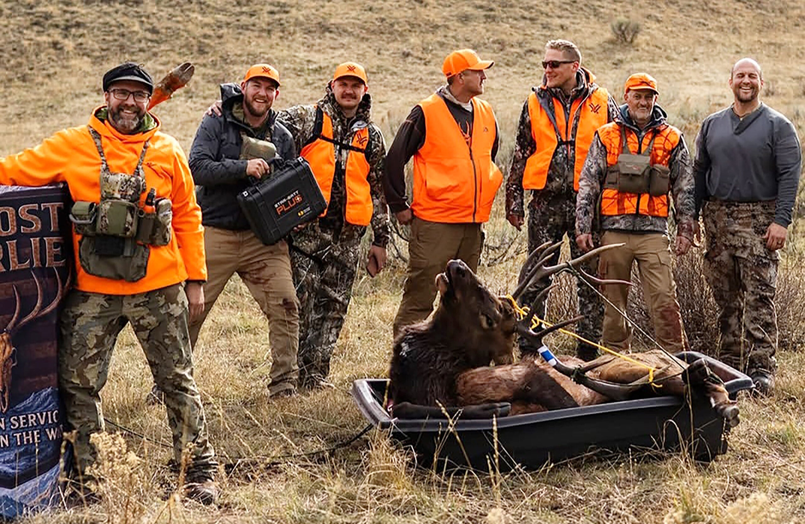 A group of hunters in orange and camouflage pose outdoors with hunting gear beside a sled carrying a large elk.