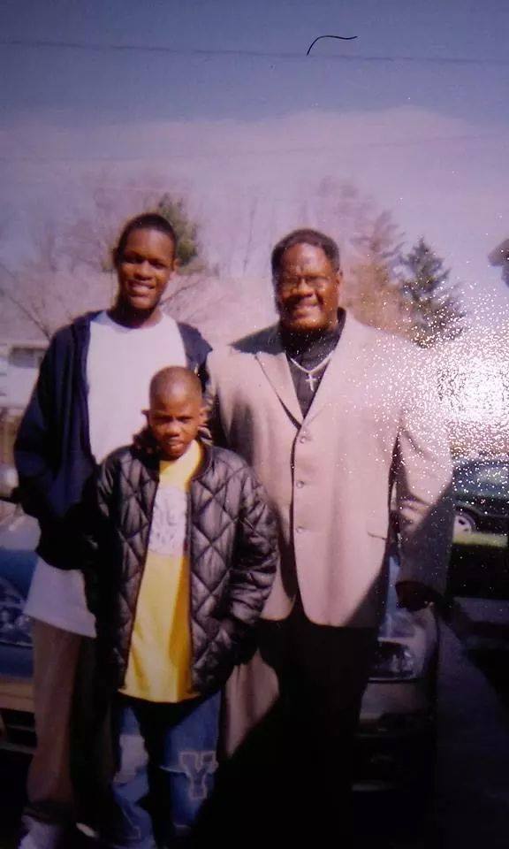 Three people, two adults and one child, stand together outdoors in daylight, posing for a photo in front of a car with trees in the background.