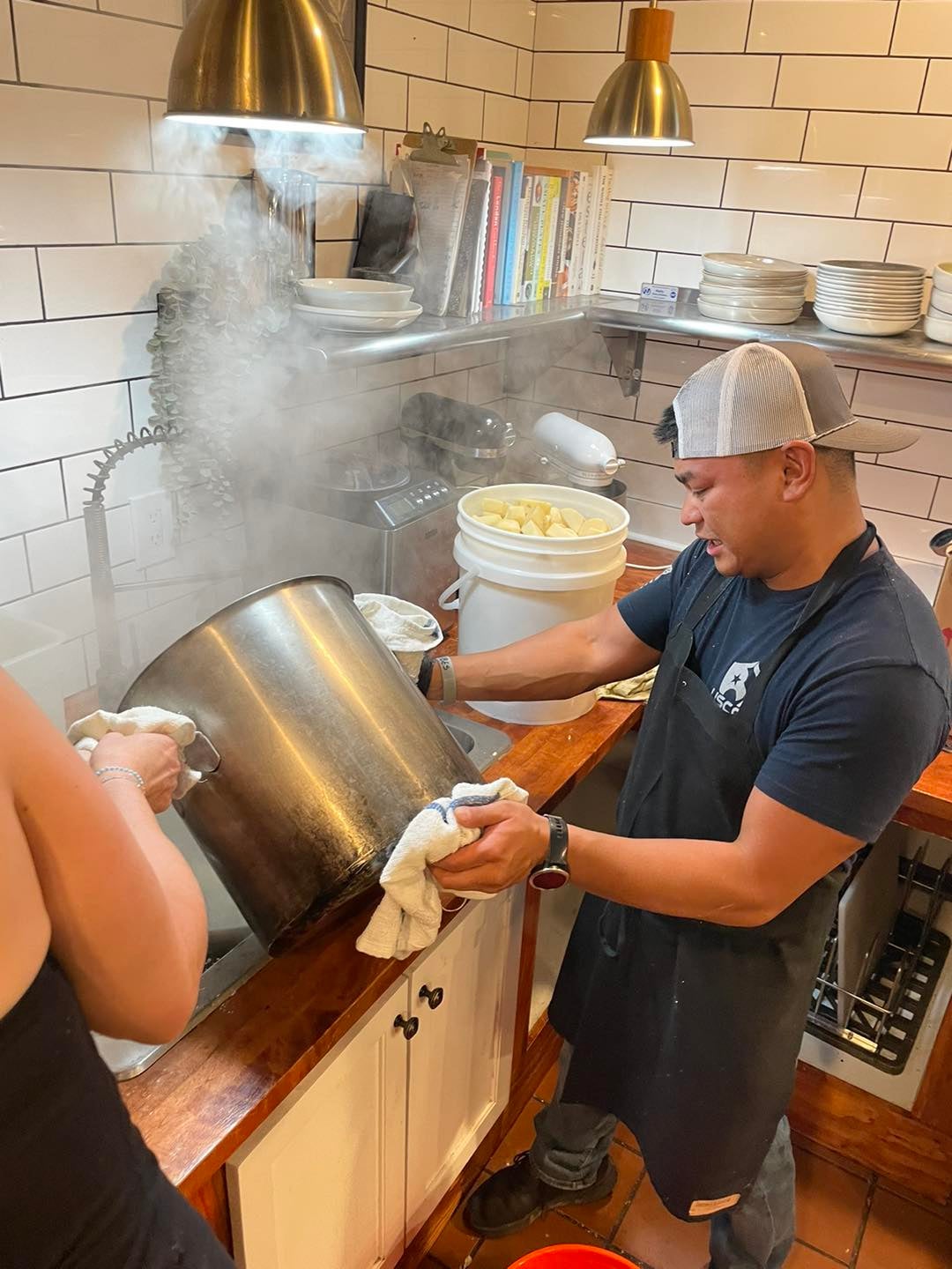 Two people in a kitchen pour steaming contents from a large pot into a sink; shelves with dishes and books are visible in the background.