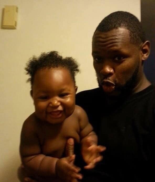 A man in a black shirt holds a smiling baby with curly hair; both are looking at the camera, and the man is making a playful facial expression.