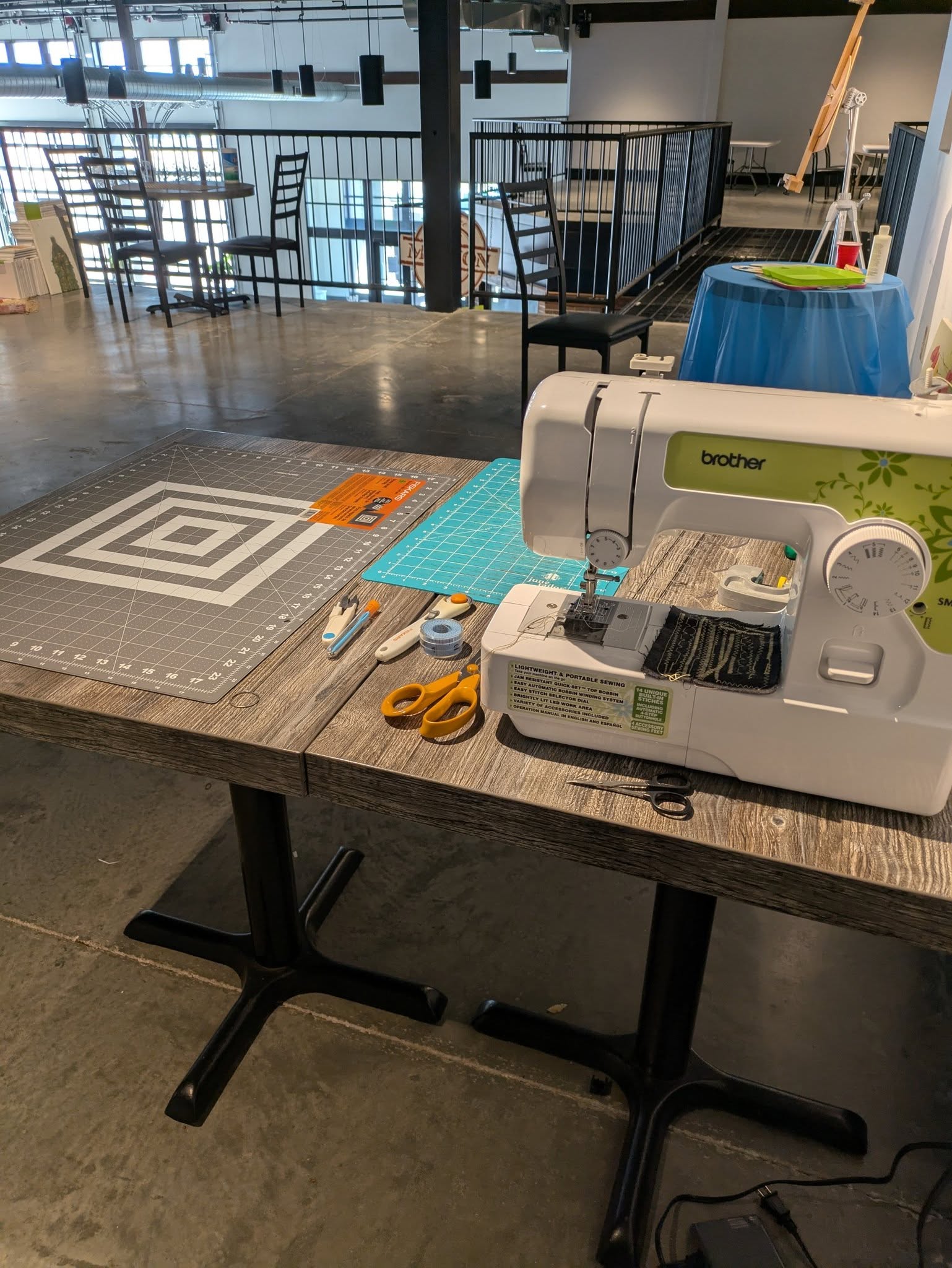 A sewing machine, cutting mats, scissors, rotary cutters, and sewing tools are arranged on a table in a spacious, modern workspace.