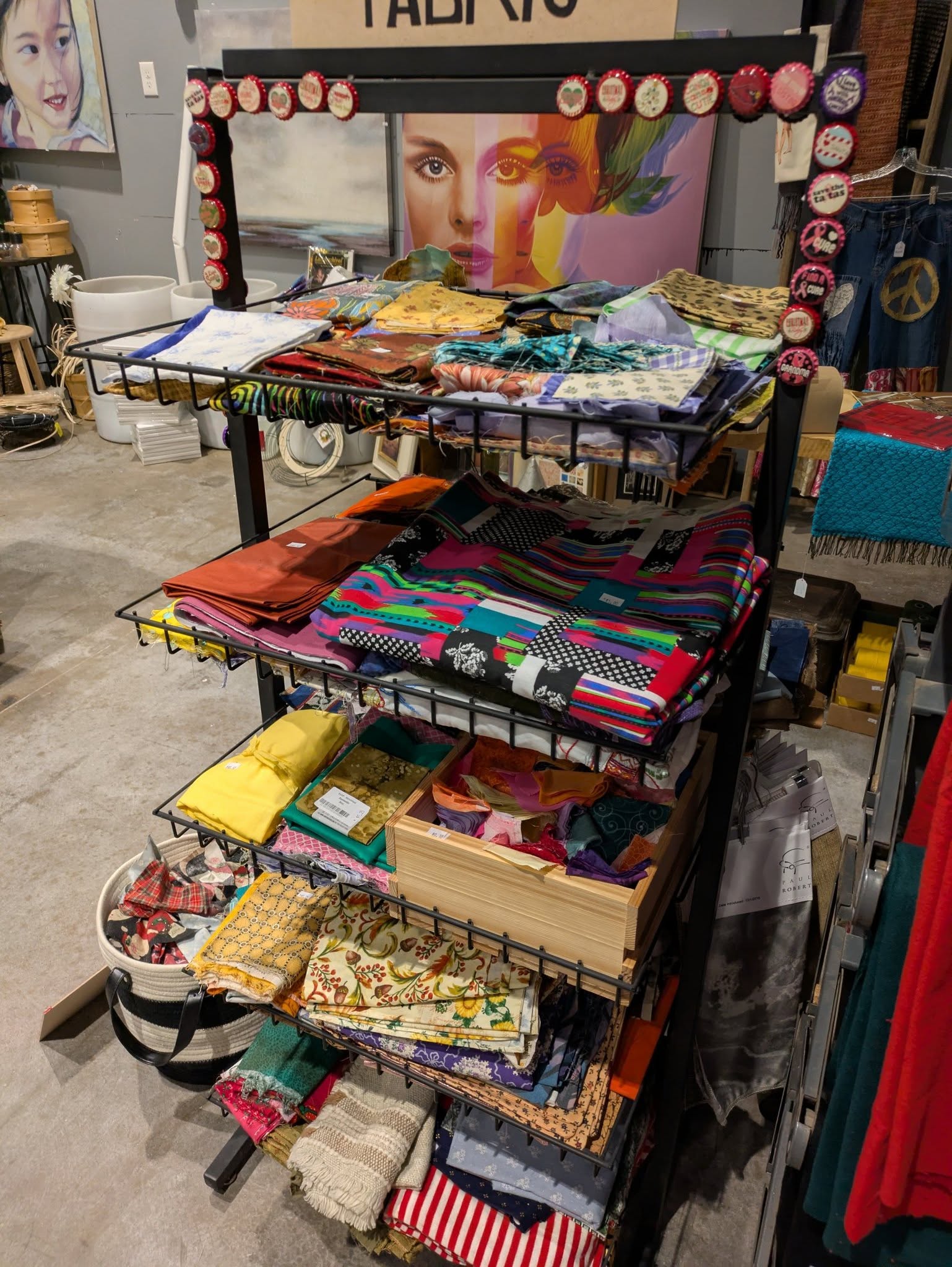 A metal display rack holds colorful folded fabric pieces and sewing supplies in a craft store, with art and textiles visible in the background.