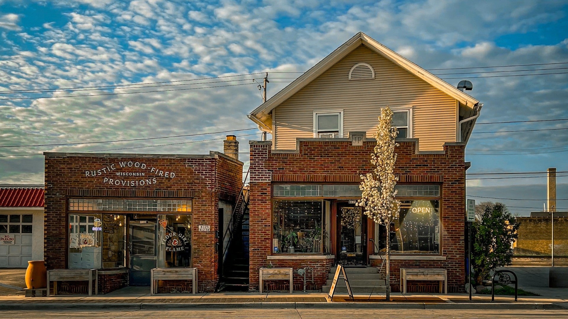 A two-story brick building with a store called Rustic Wood Fired Provisions on the left and a shop with an Open sign on the right, under a partly cloudy sky.