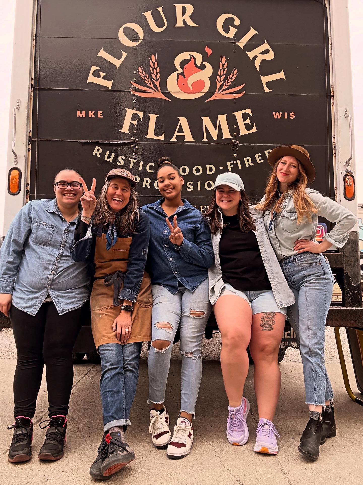 Five women pose and smile in front of a food truck that reads Flour Girl & Flame, all wearing casual denim and comfortable clothing.