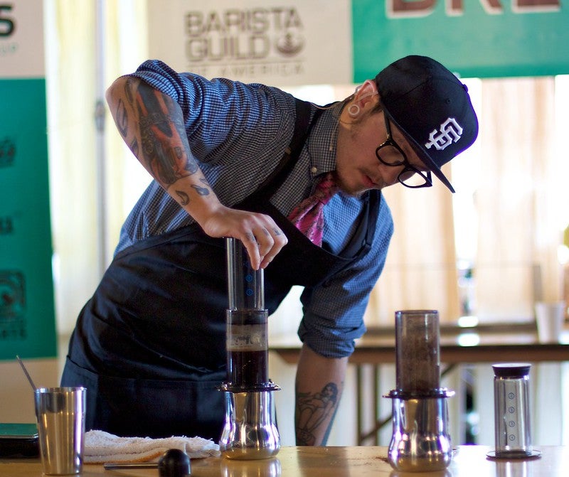 A barista wearing glasses, a hat, and tattoos is preparing coffee using an AeroPress at a competition or event.