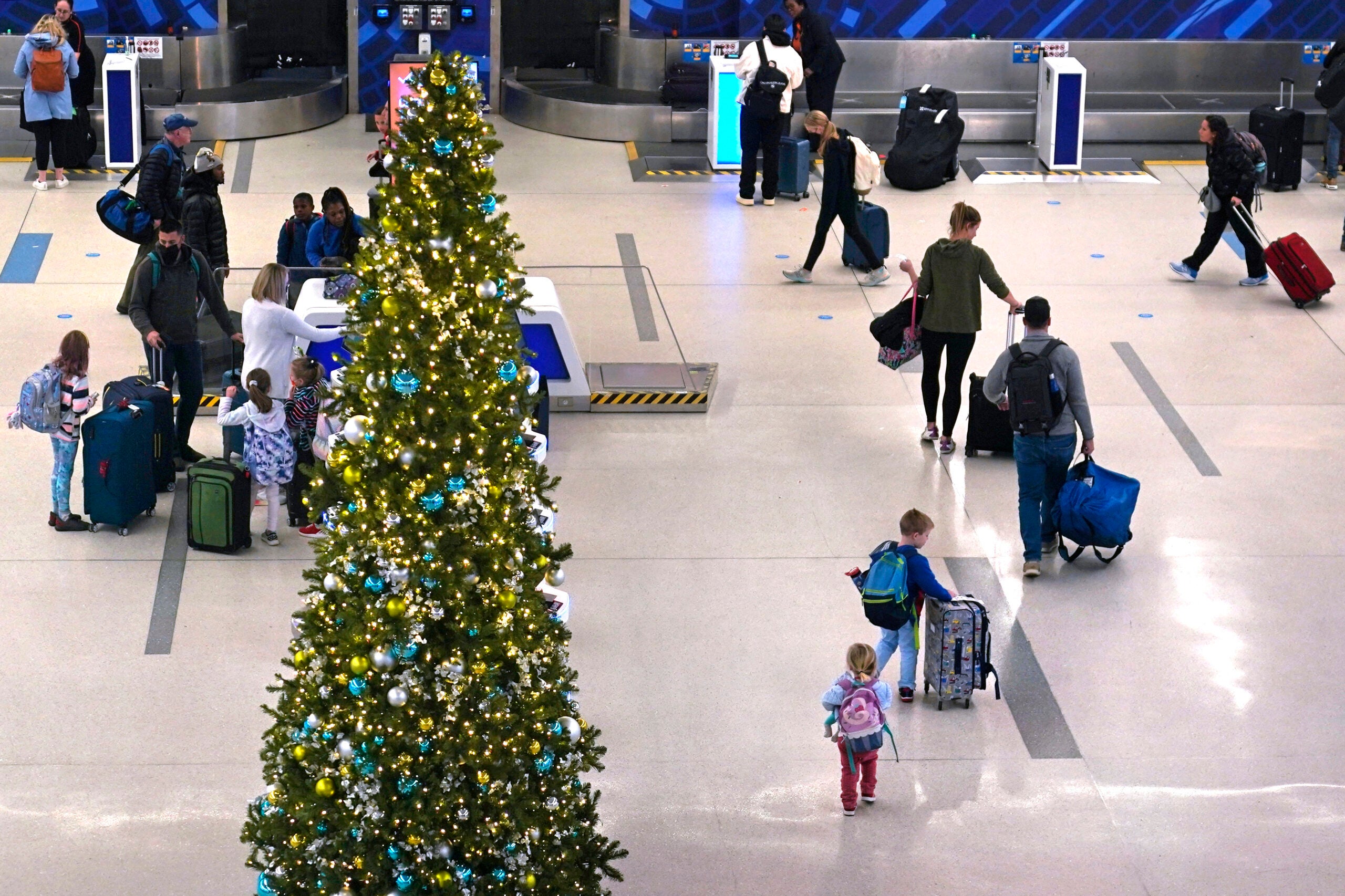 Travelers with luggage walk near a decorated Christmas tree in an airport terminal, with baggage claim stations in the background.