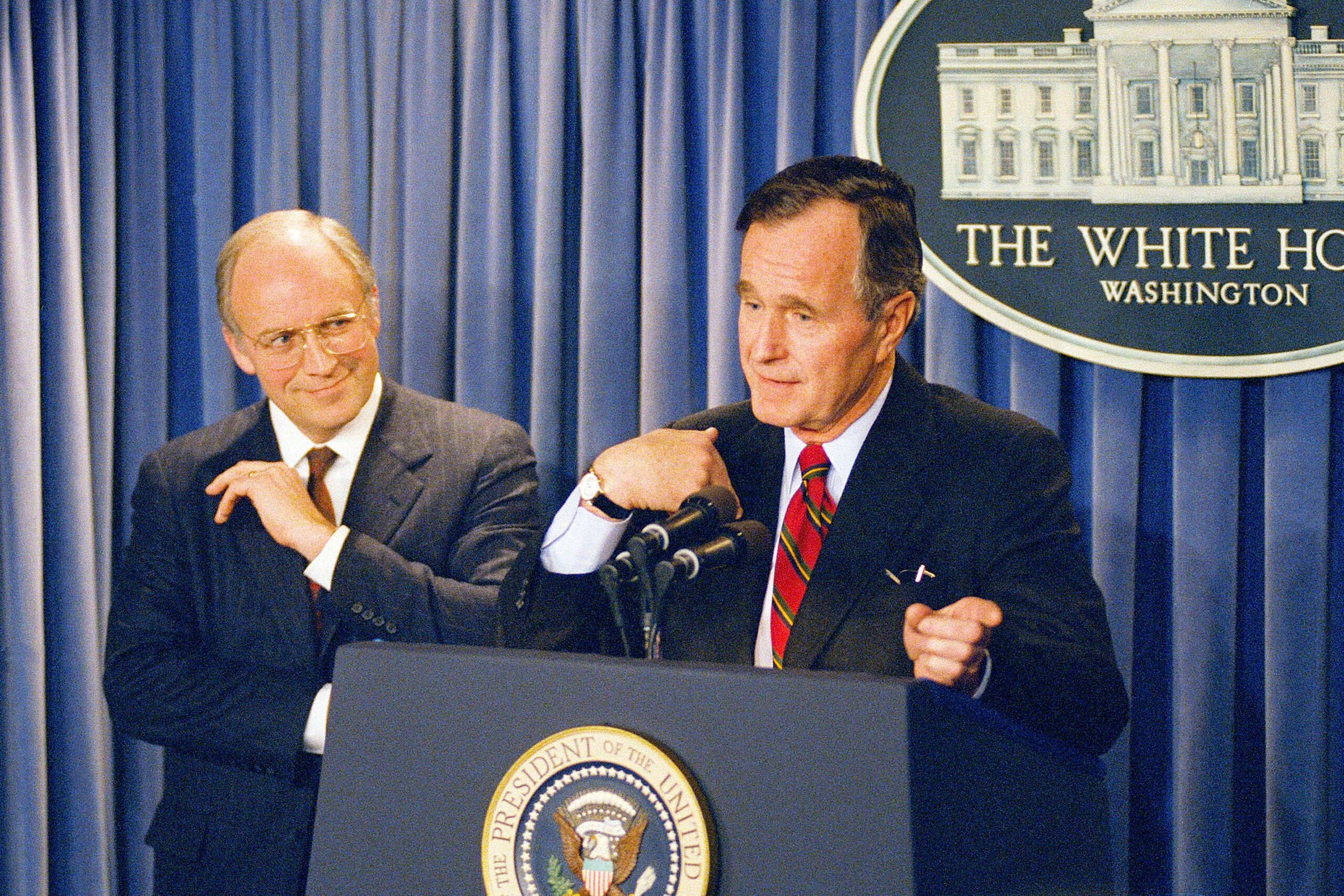 Two men in suits stand at a podium with the U.S. presidential seal; one speaks into microphones, the other stands to the side, both in front of a White House sign.