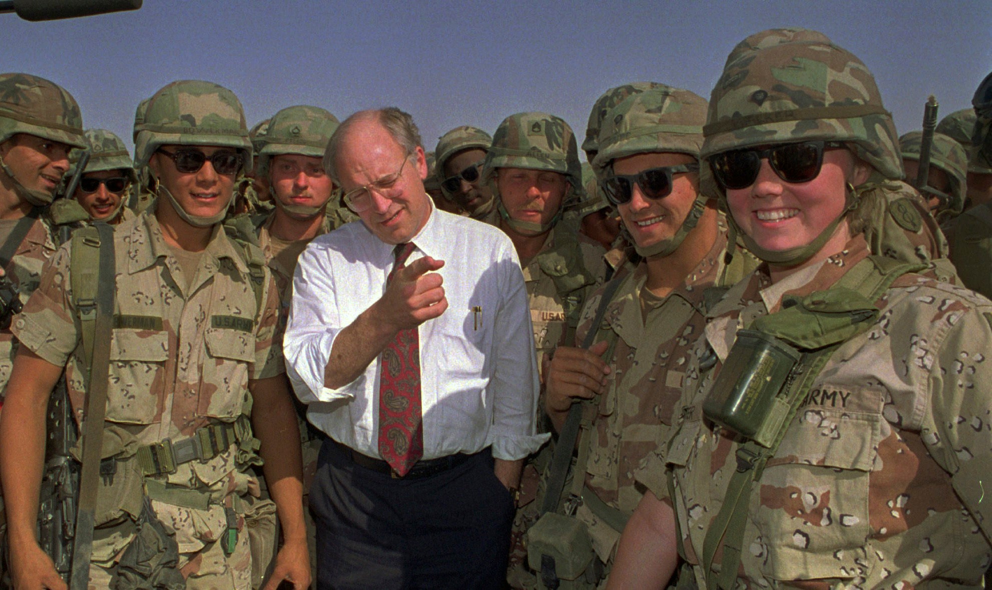 A man in a white shirt and tie stands among smiling soldiers in military uniforms and helmets under a clear sky.