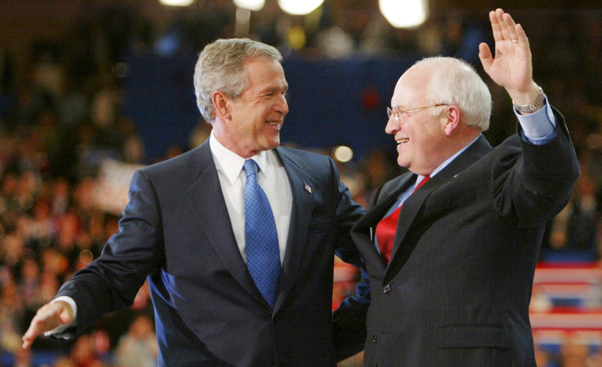 Two men in suits, one with a blue tie and the other with a red tie, smile and gesture on stage in front of a blurred audience.