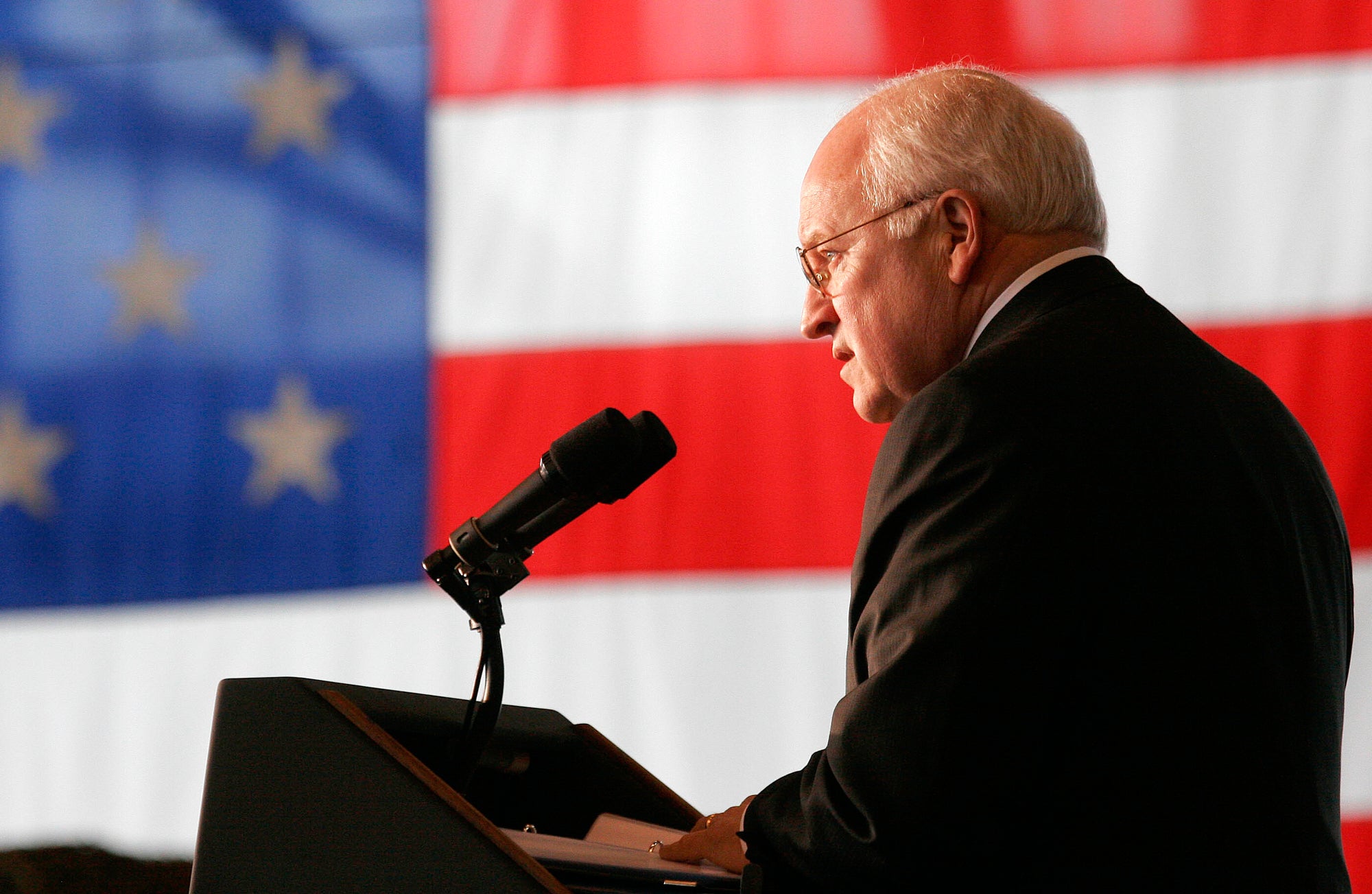 A man in a suit speaks into a microphone at a podium with a large American flag in the background.