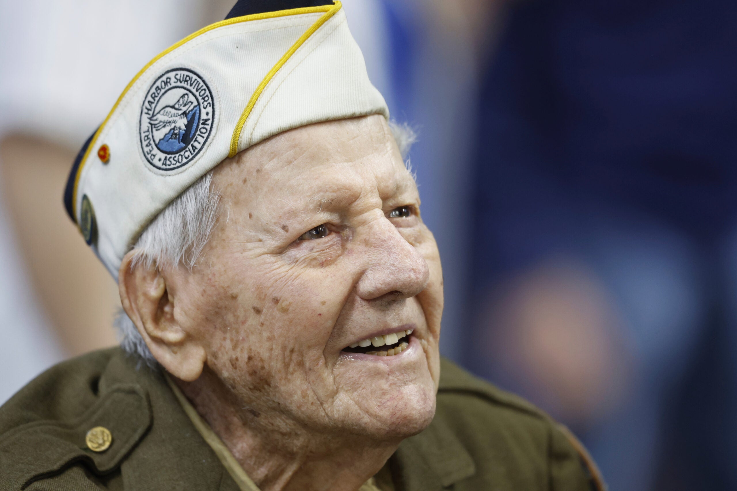 Elderly man wearing a military uniform and a white cap with a Pearl Harbor Survivors Association patch, smiling and looking slightly upward.