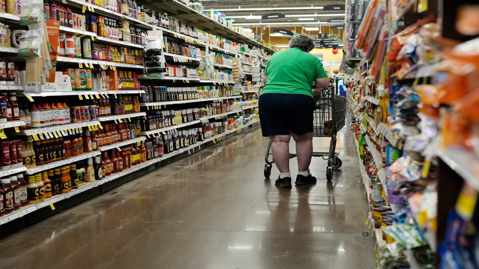 A person in a green shirt and blue shorts pushes a shopping cart down a grocery store aisle lined with various food products.