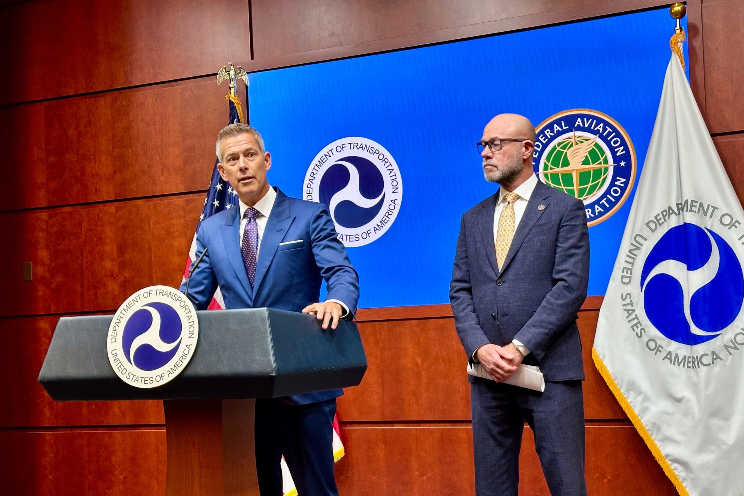 Two men in suits stand at a podium with Department of Transportation and FAA logos, speaking at a press conference in front of official flags and a blue background.