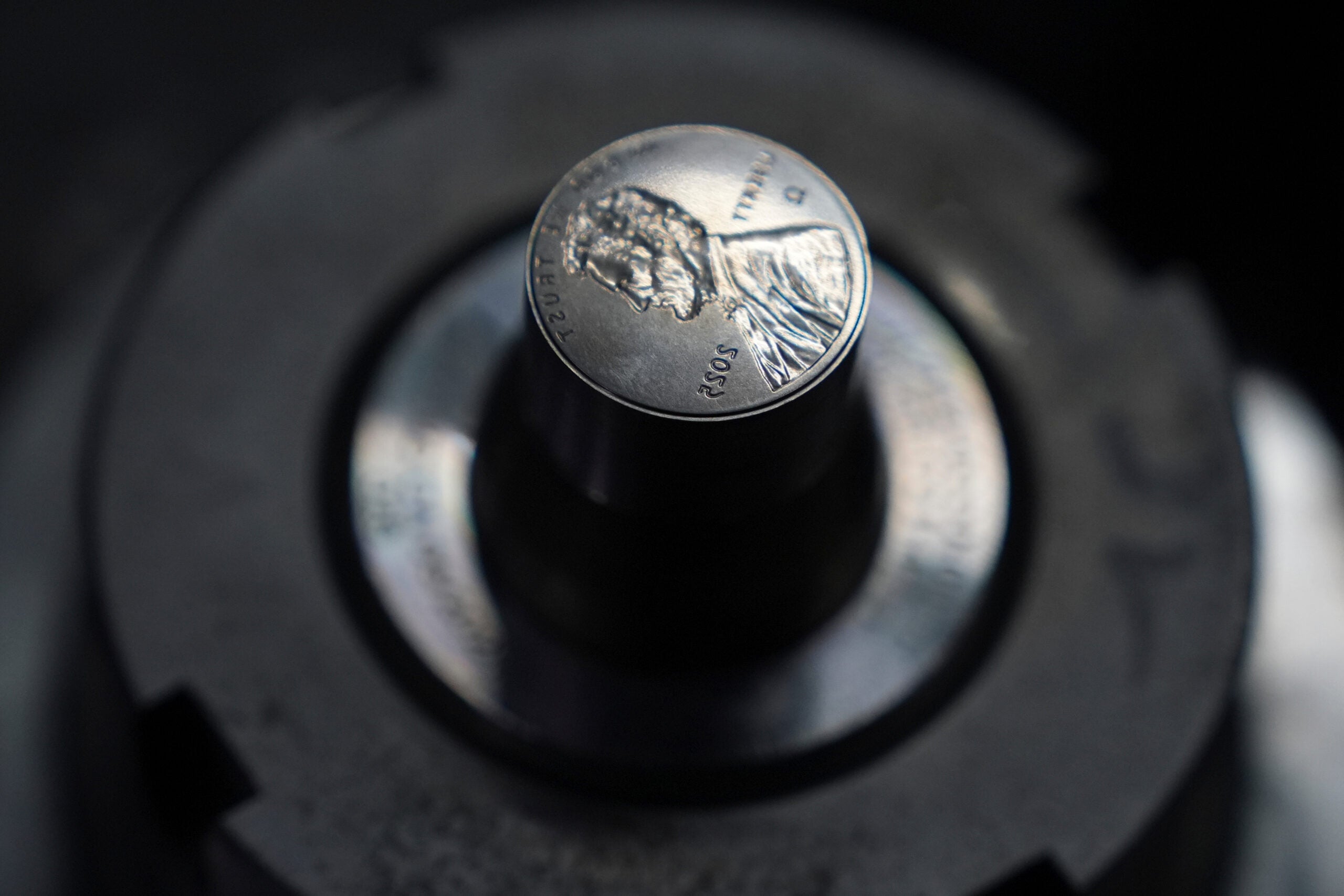 Close-up of a metal coin, possibly a penny, positioned upright on a metallic machine part, with the background blurred.