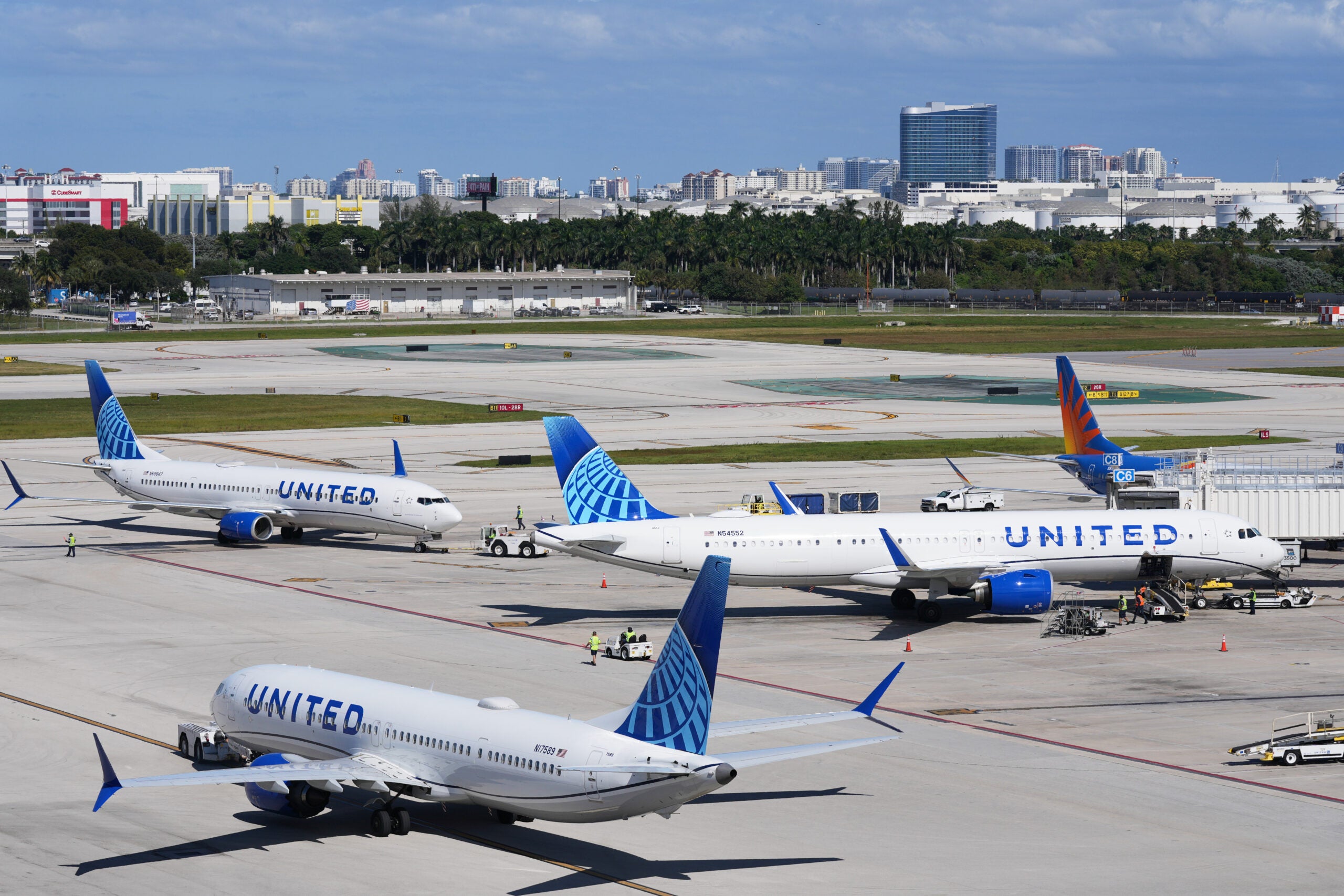 Several United Airlines planes are parked and taxiing at an airport terminal on a clear day, with a city skyline visible in the background.