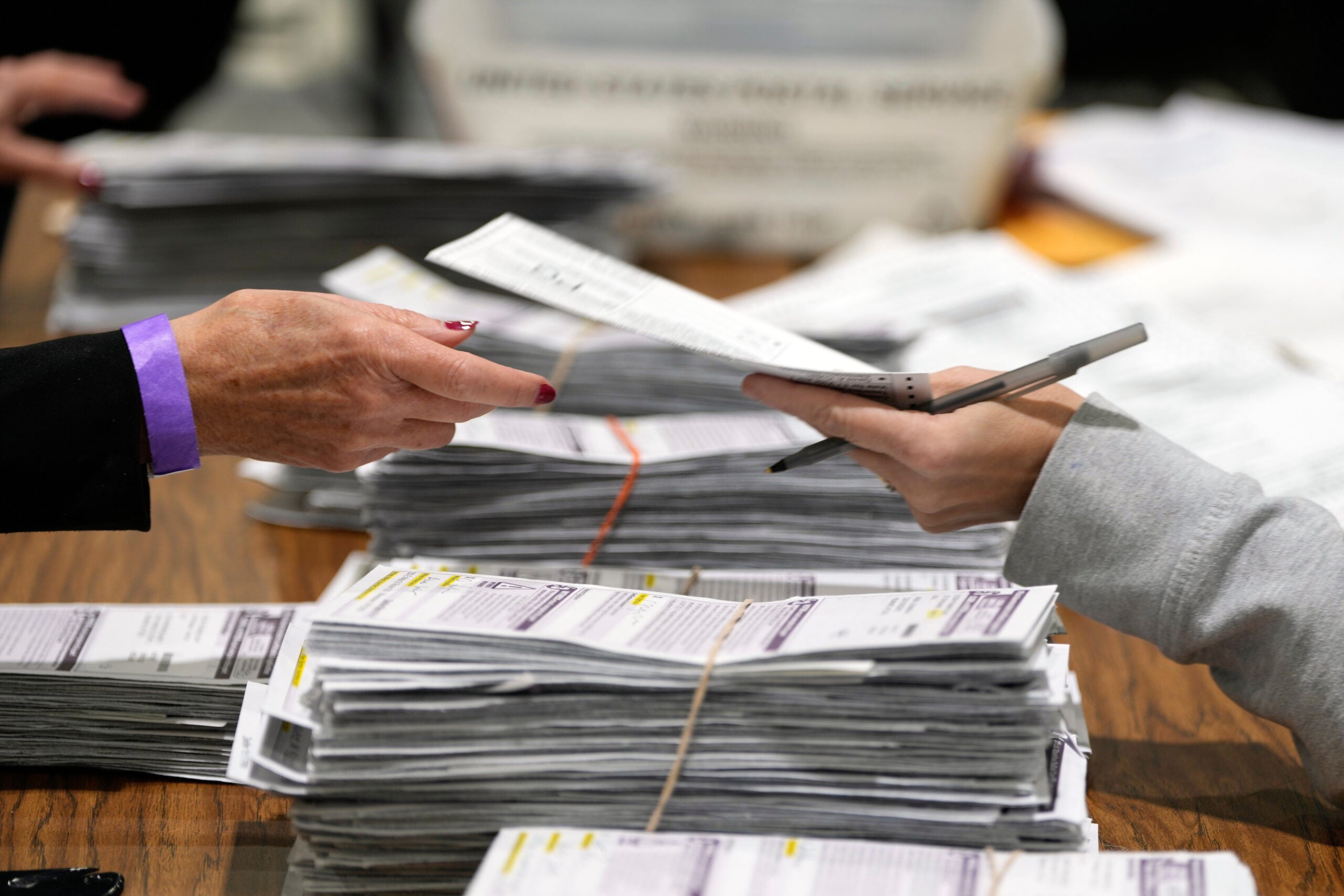 Two people exchange paperwork over stacks of documents on a table, with additional papers and a container visible in the background.