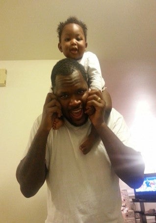 An adult man in a white shirt holds a child on his shoulders indoors. Both are smiling and facing the camera.