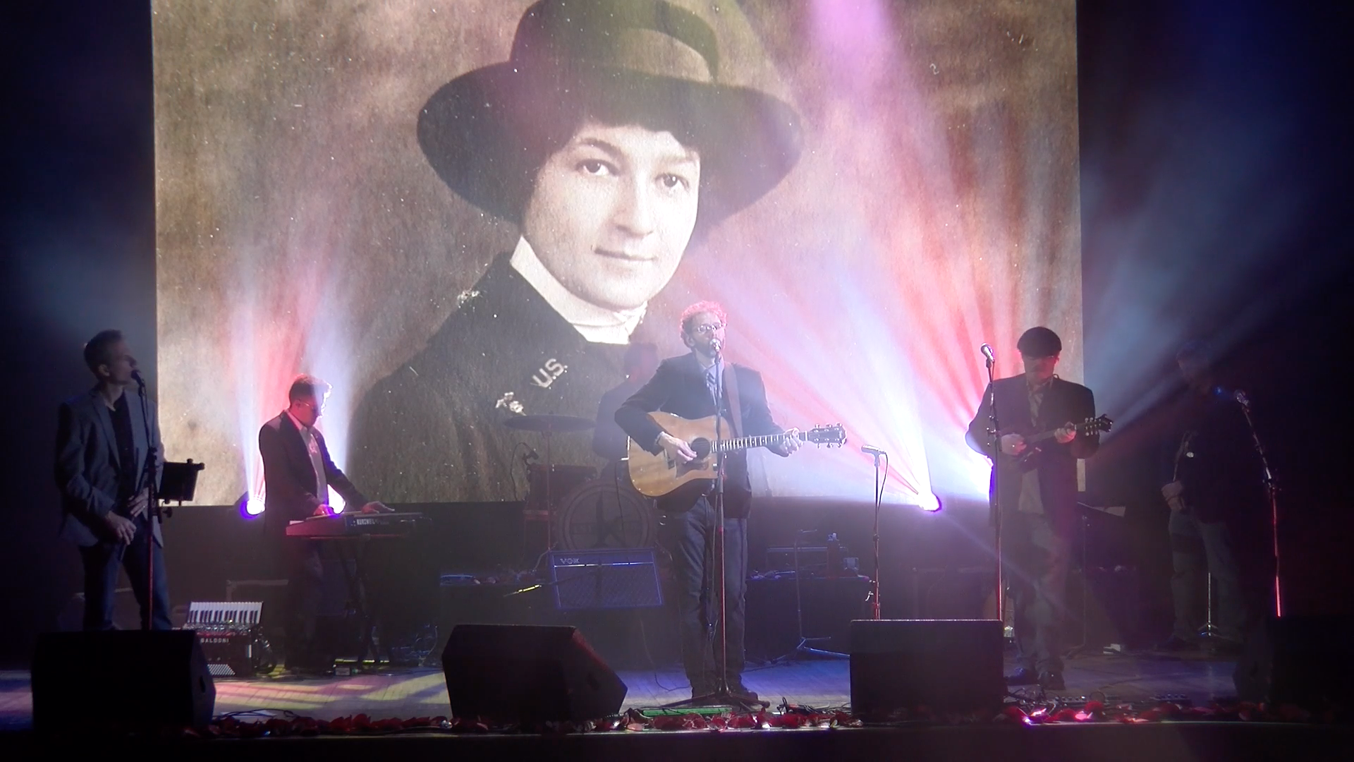 A band performs on stage with colorful lights, while a large vintage photograph of a woman in uniform is projected in the background.