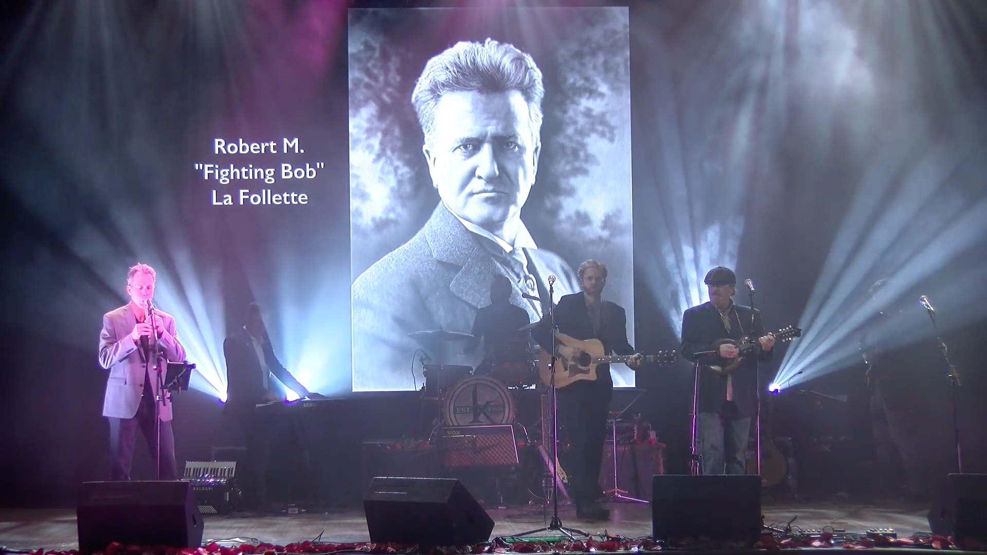 Three musicians perform on stage in front of a large black-and-white portrait labeled Robert M. Fighting Bob La Follette, with dramatic stage lighting and instruments visible.
