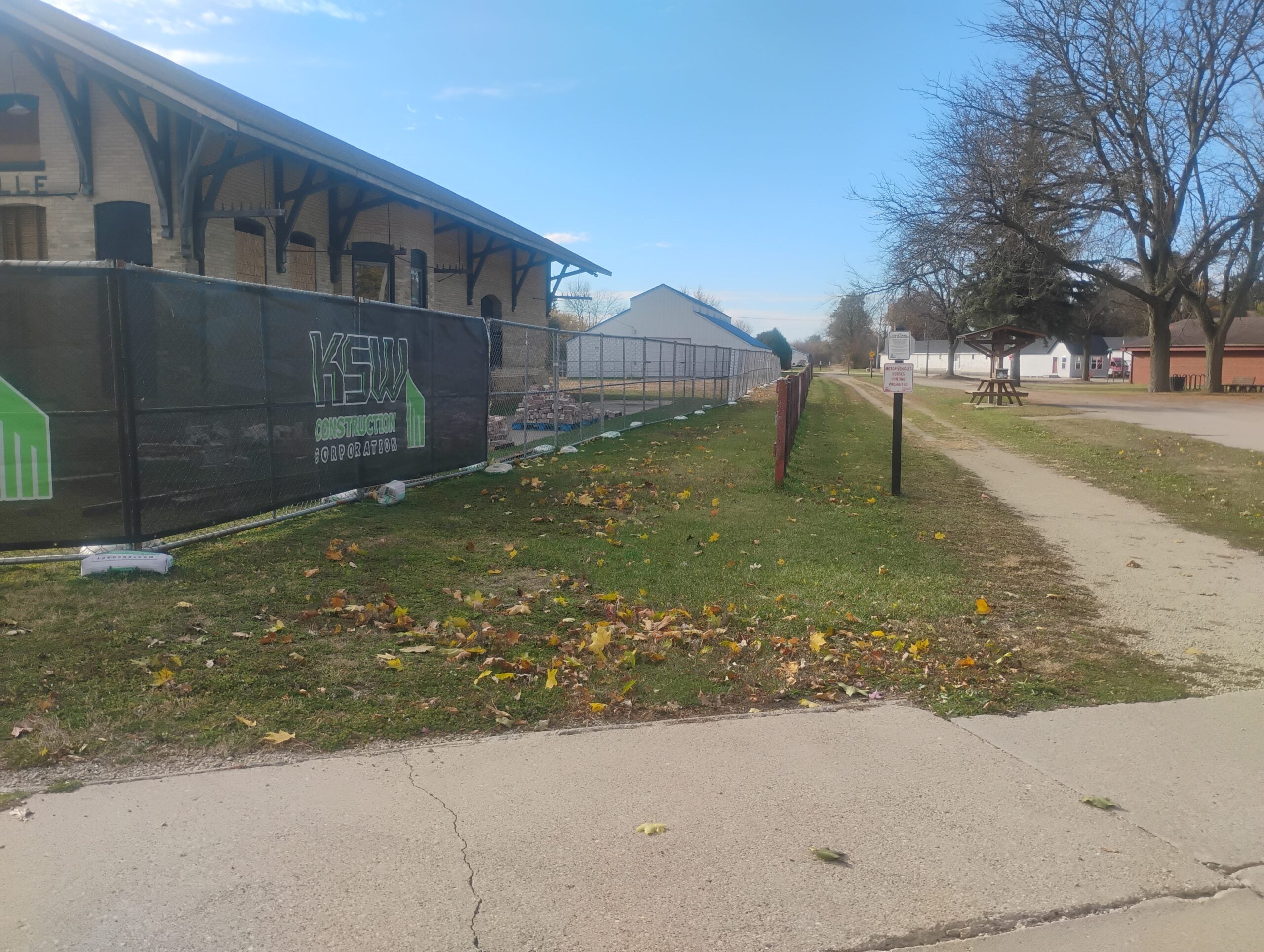A fenced construction site runs alongside a sidewalk and grassy area with scattered autumn leaves; trees and buildings are visible in the background.