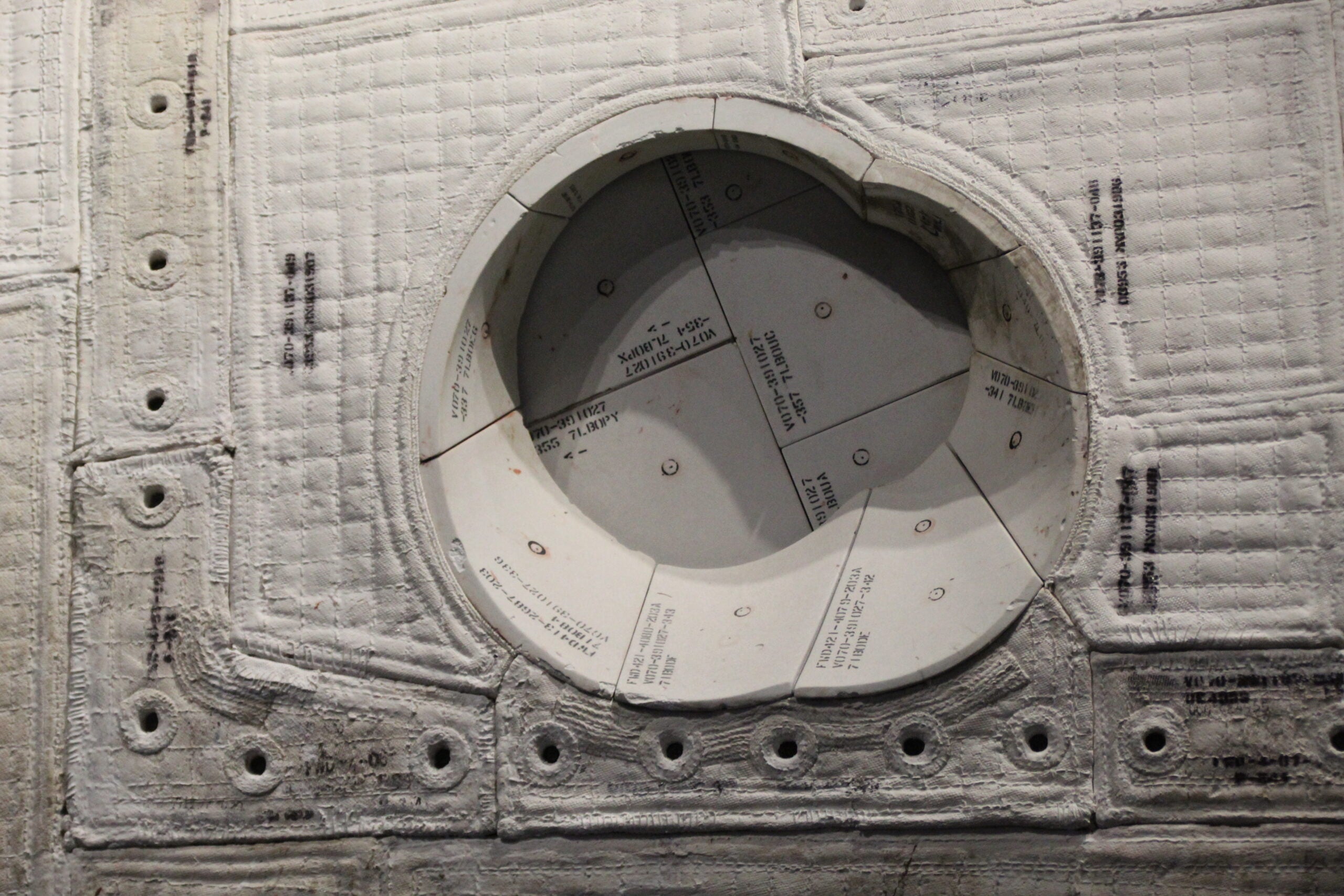 Close-up of a circular hatch opening on a spacecraft covered with white thermal insulation tiles, showing bolts and labeled segments. Photo courtesy of Wright