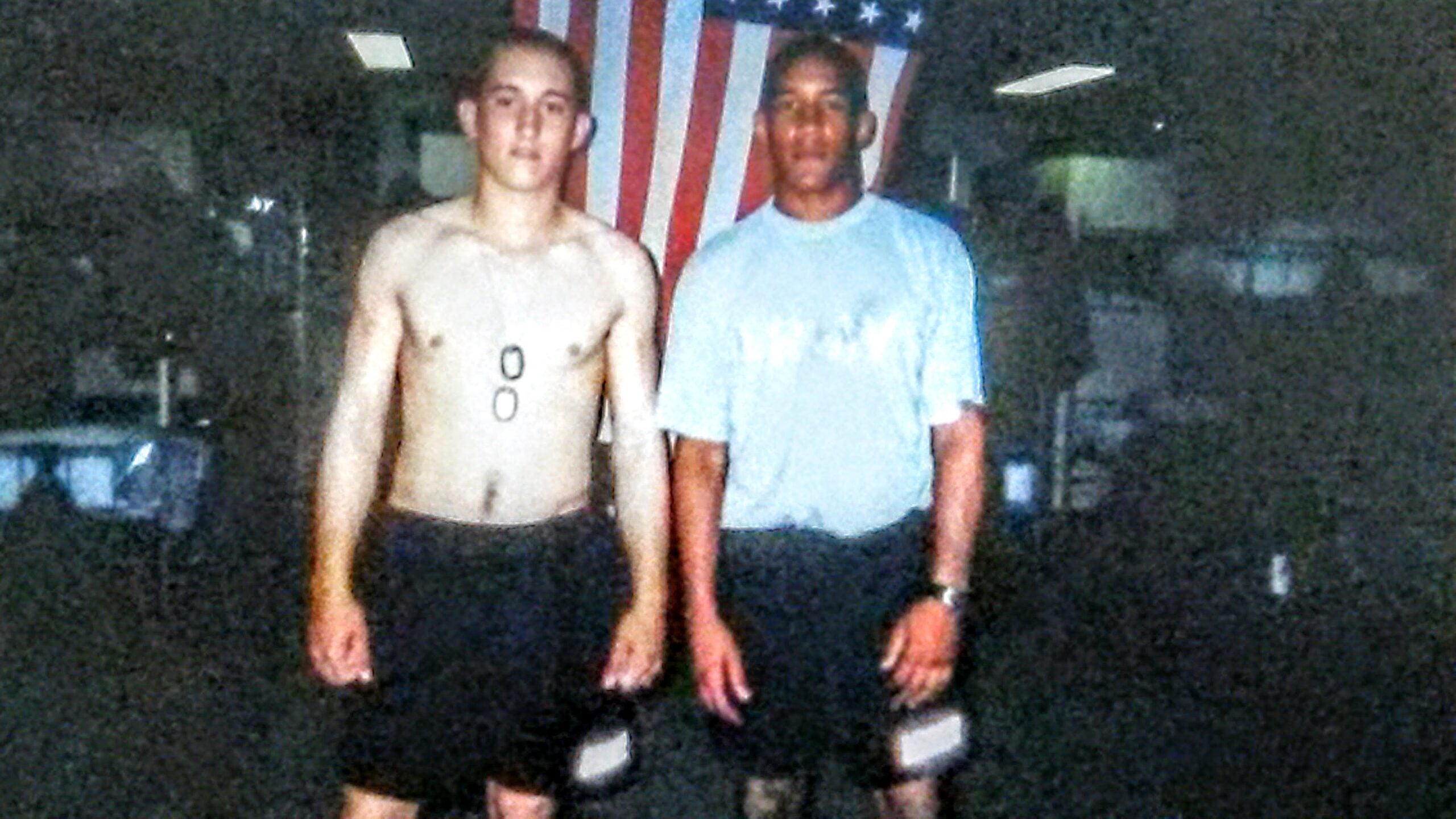 Two men stand side by side in shorts, one shirtless with dog tags, in front of an American flag inside a gym or training facility.